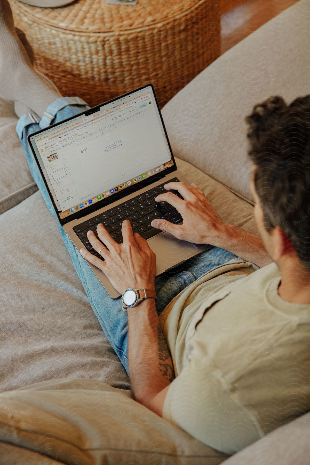 A man sits on a couch, working on his laptop, with a wicker basket in the background.