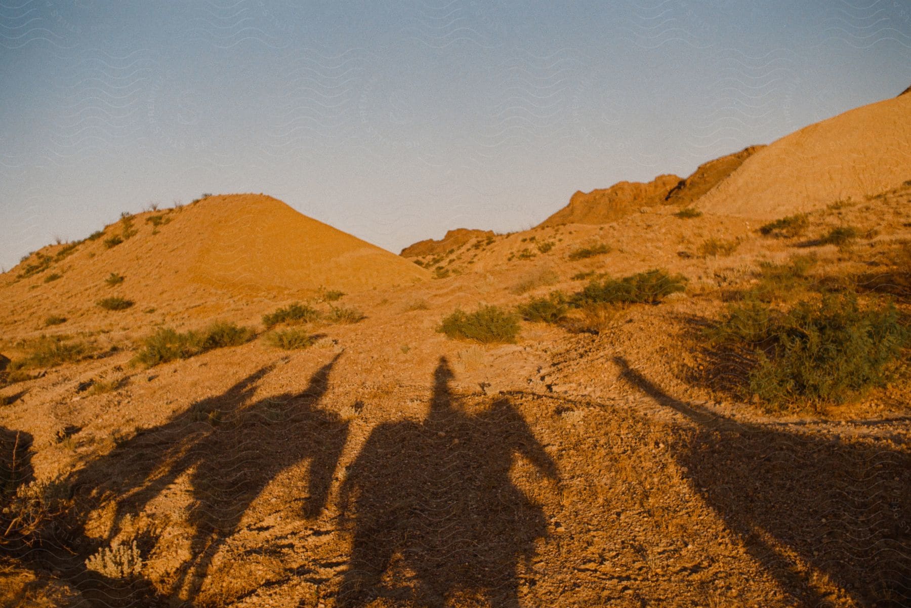 The shadows of several people riding on horseback are cast upon the desert ground.