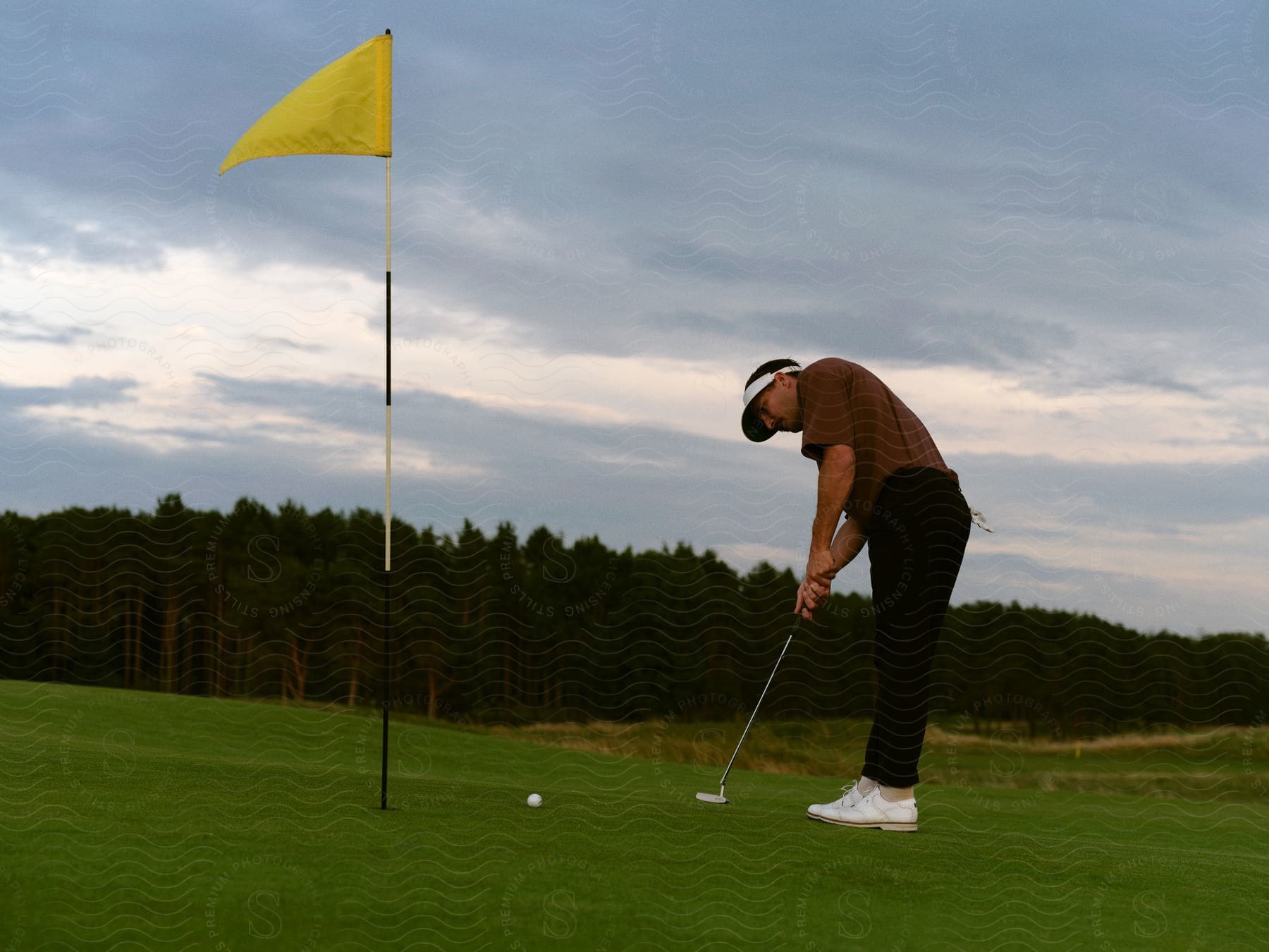 A golfer putts his ball towards the flagpole in the hole on a cloudy day, with a forest in the background.