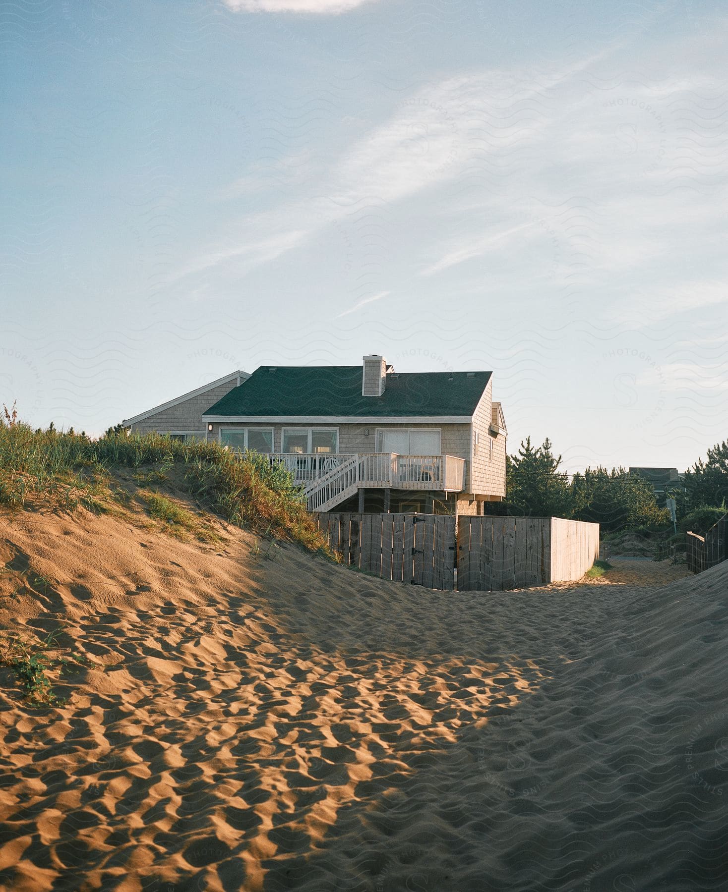 A sandy path with footprints leads to a beach house hidden behind a sand dune, with a wooden fence surrounding it.