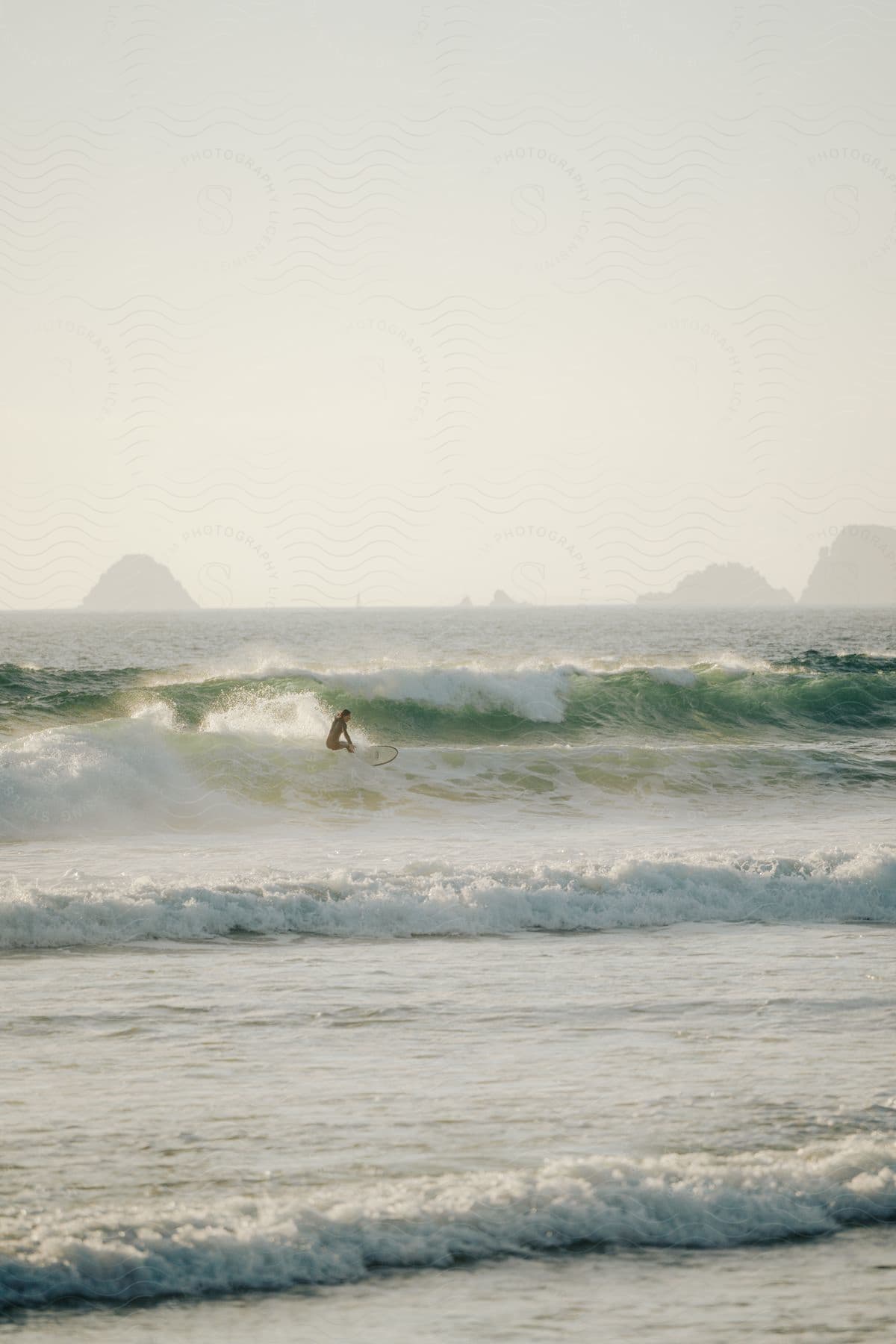 A surfer rides a wave into shore with tall rock formations in the distance under a hazy sky