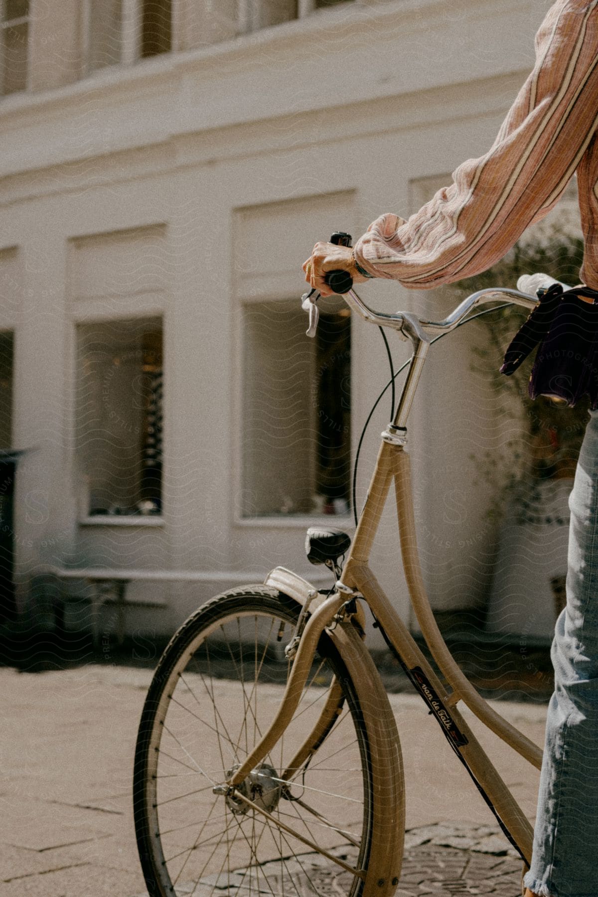 A person in long sleeves stands next to a bike on a street in front of a building.