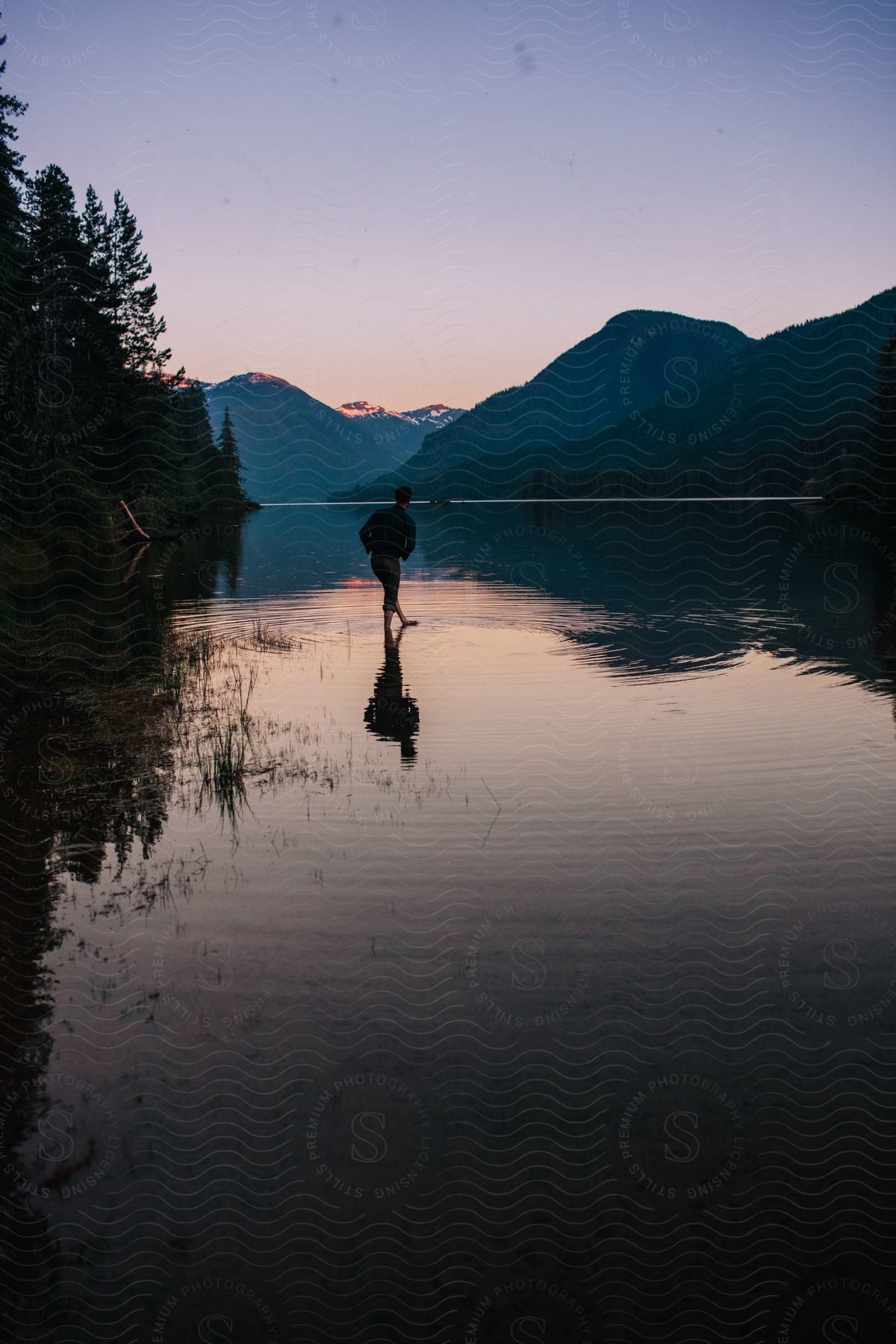 Man walking in serene lake is silhouetted against the mountain range and sky at sunset