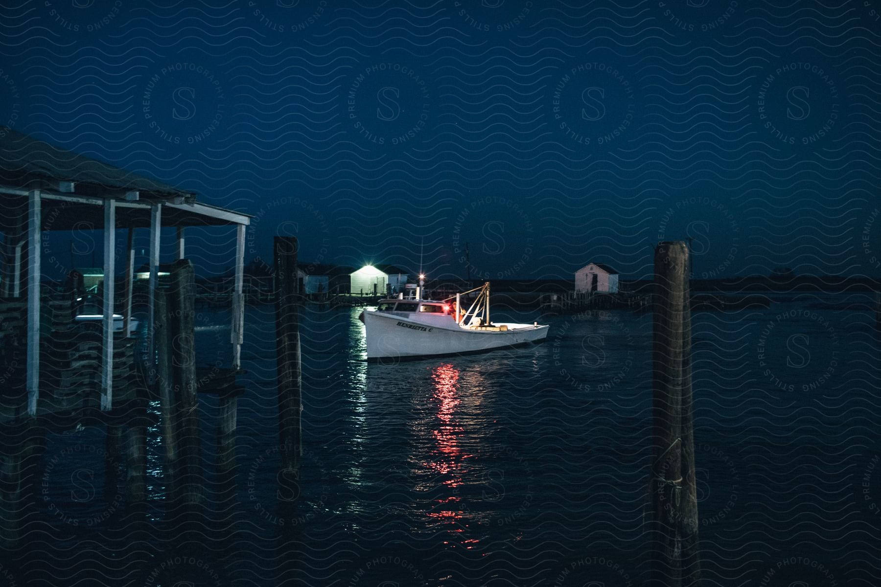 A small fishing boat, its lights glowing in the night, returns to port with its catch, the pier and storage units looming behind it.