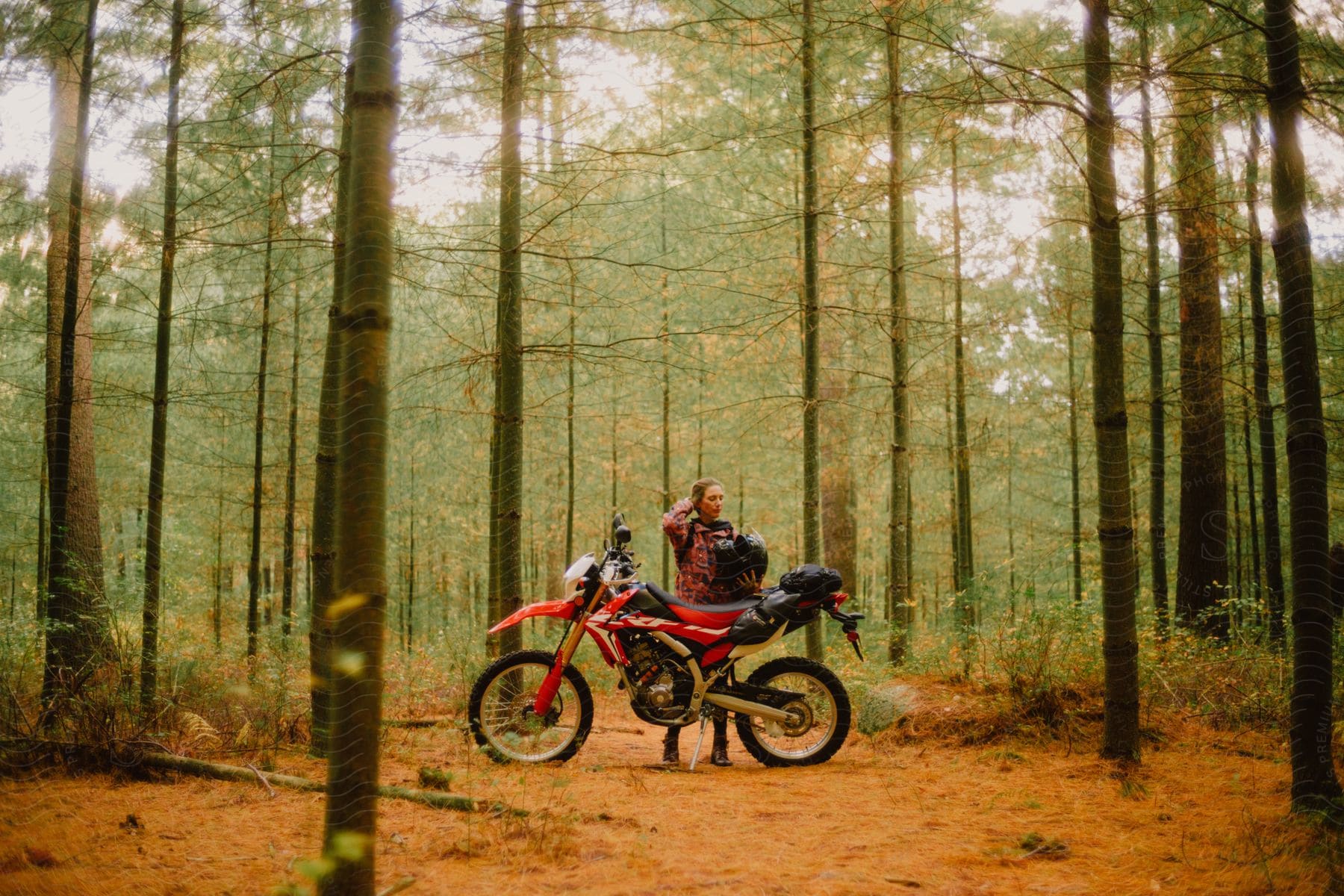 A woman in sportswear, helmet in hand, poses next to her red motorbike on a orange dirt road in the woods, surrounded by autumn foliage.