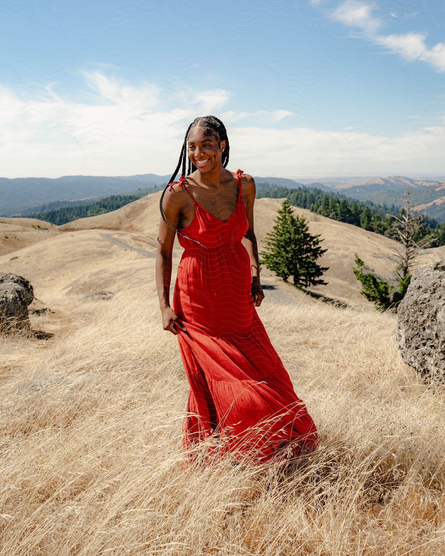 A woman wearing a long red dress smiles as she stands on a hilltop covered in tall dry grass with mountains in the distance