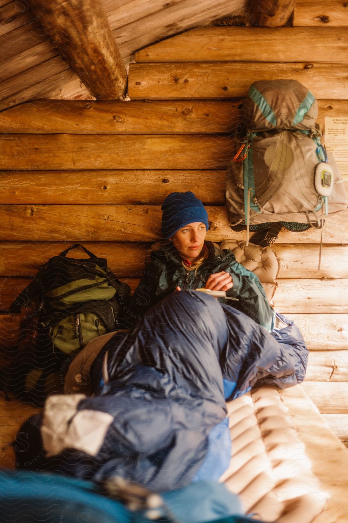 A woman in winter clothes rests in a sleeping bag on a bed in a log cabin.