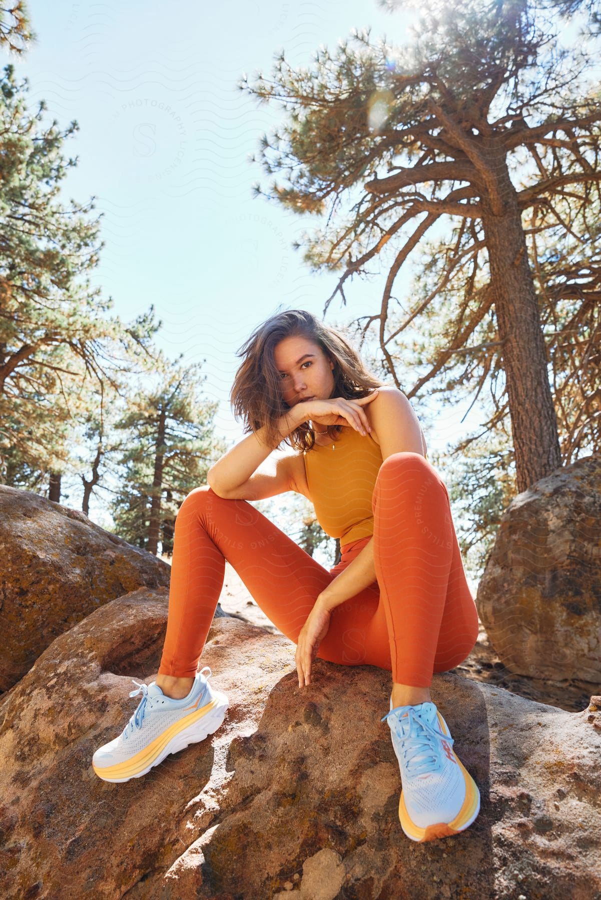 A teenage girl is sitting on boulders near trees with an elbow on her knee and hand in front of her chin