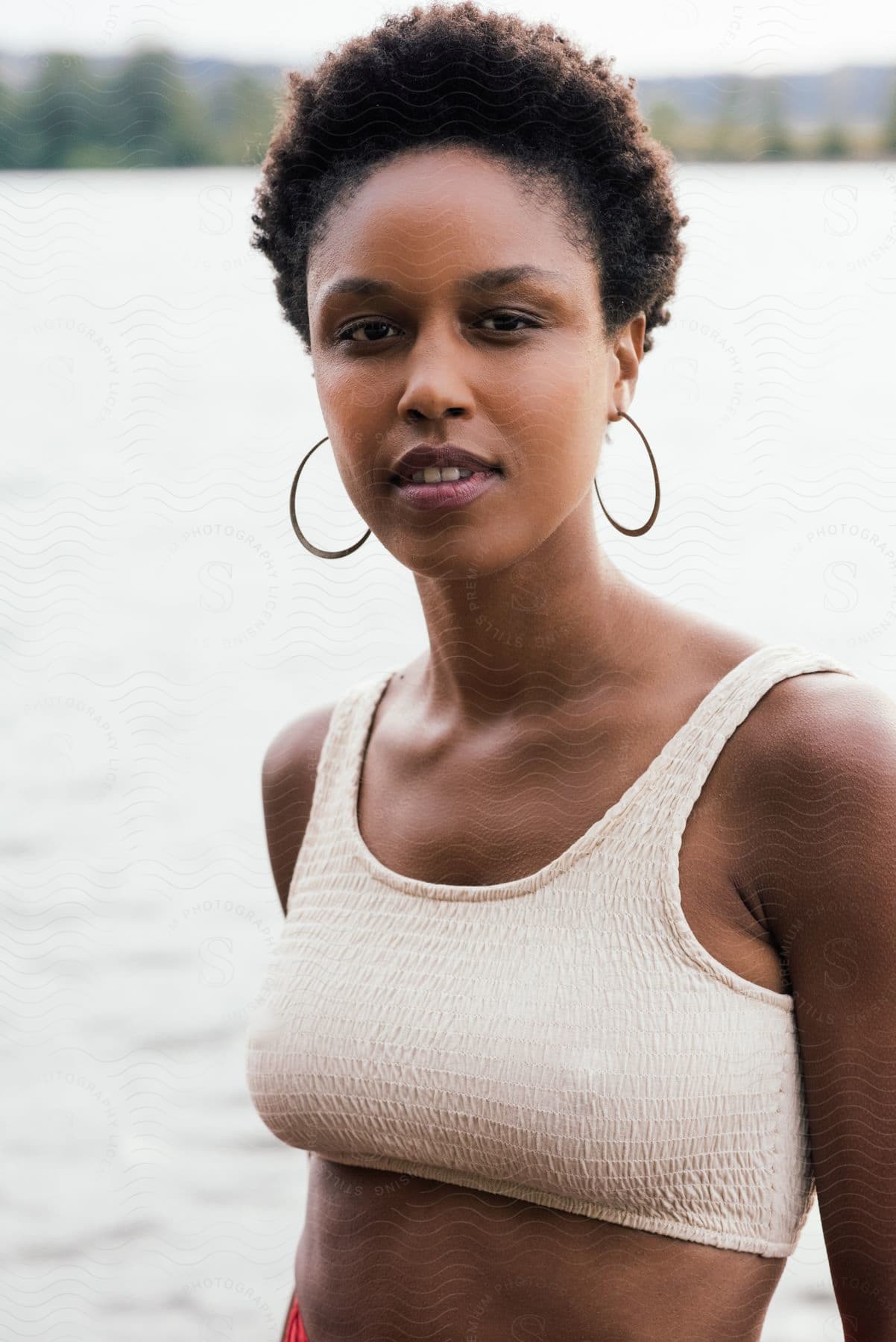 Woman with hoop earrings white top and short curly hair smiles at camera near the lake