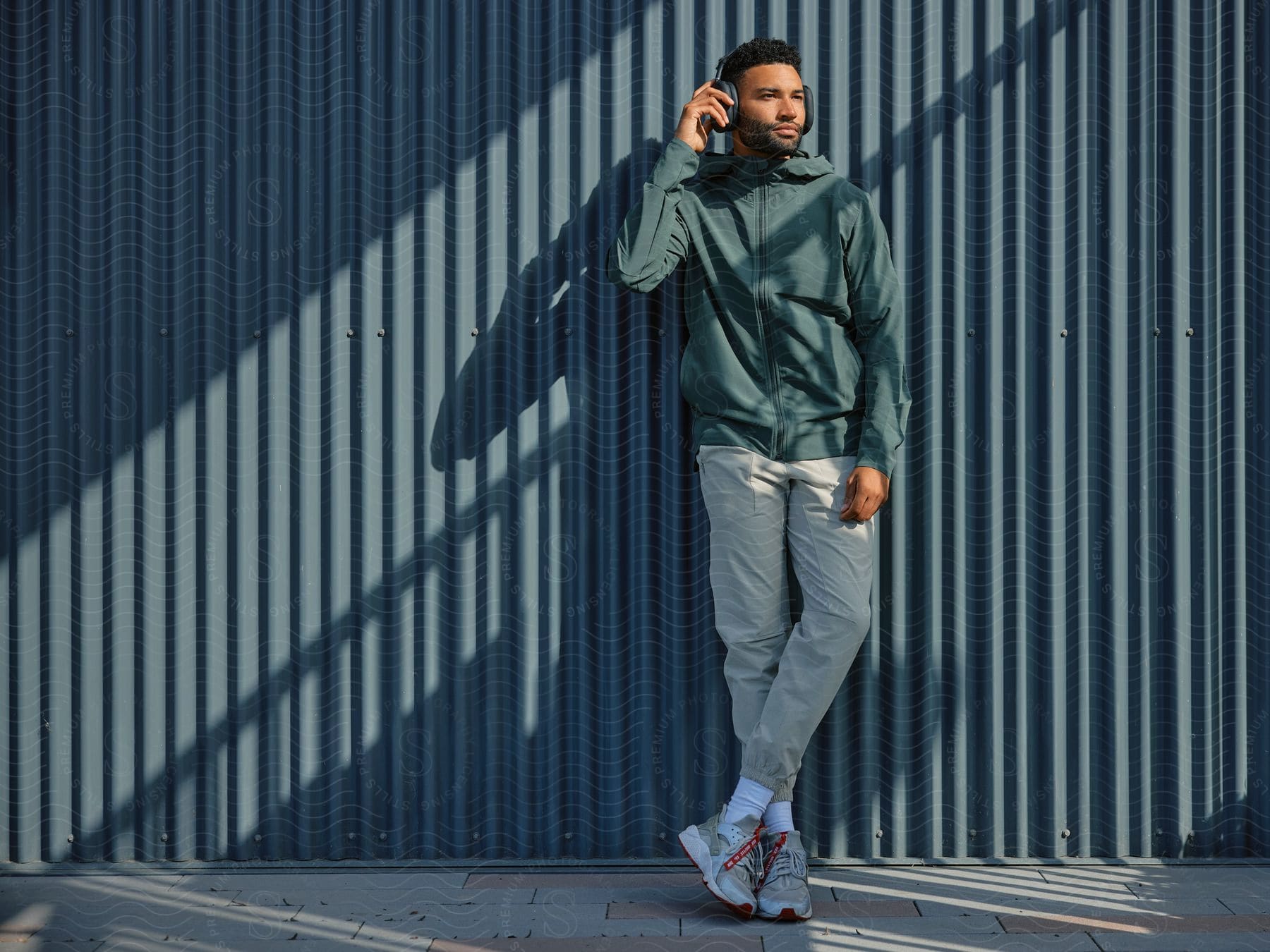 A man is leaning against a building listening to something on headphones.