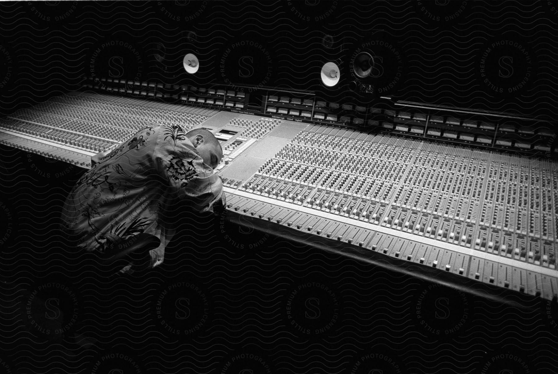 Stock photo of a young man is sleeping against a large, sound mixing board in a studio.