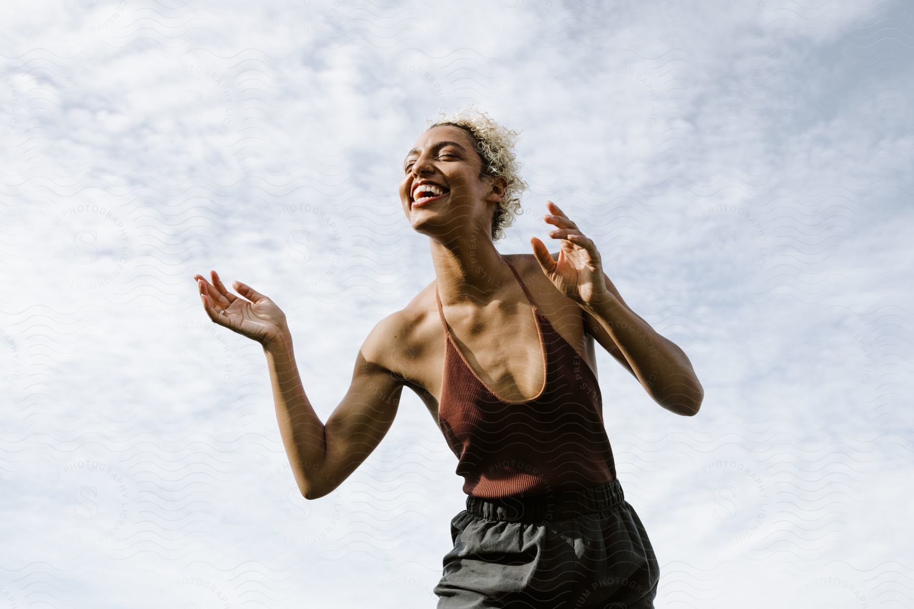 A woman smiles with her eyes closed as she stands under a cloudy sky on a sunny day.