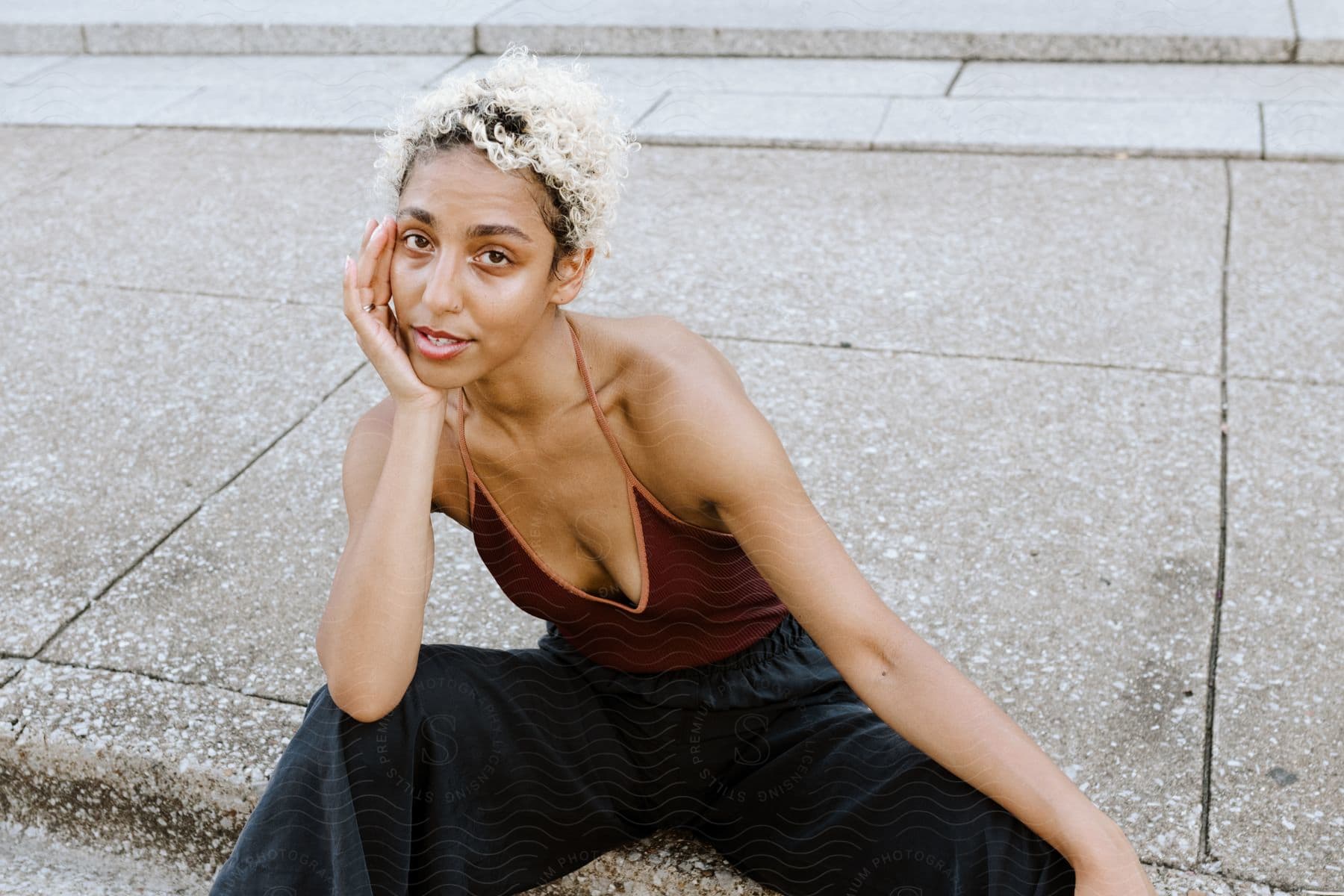 A beautiful young woman with blonde hair sits on a sidewalk while posing for the camera.