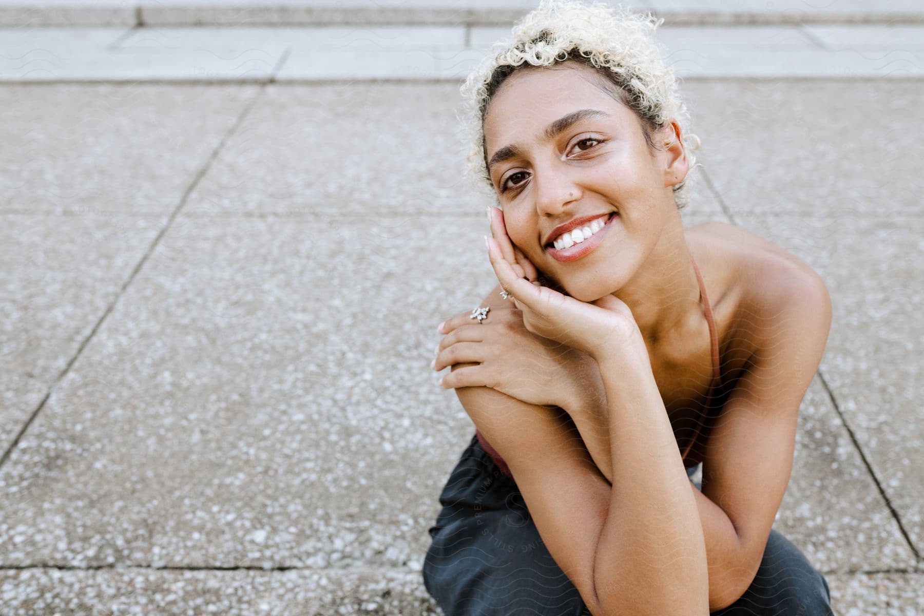 Woman smiles while crouching in sleeveless dress on sidewalk.