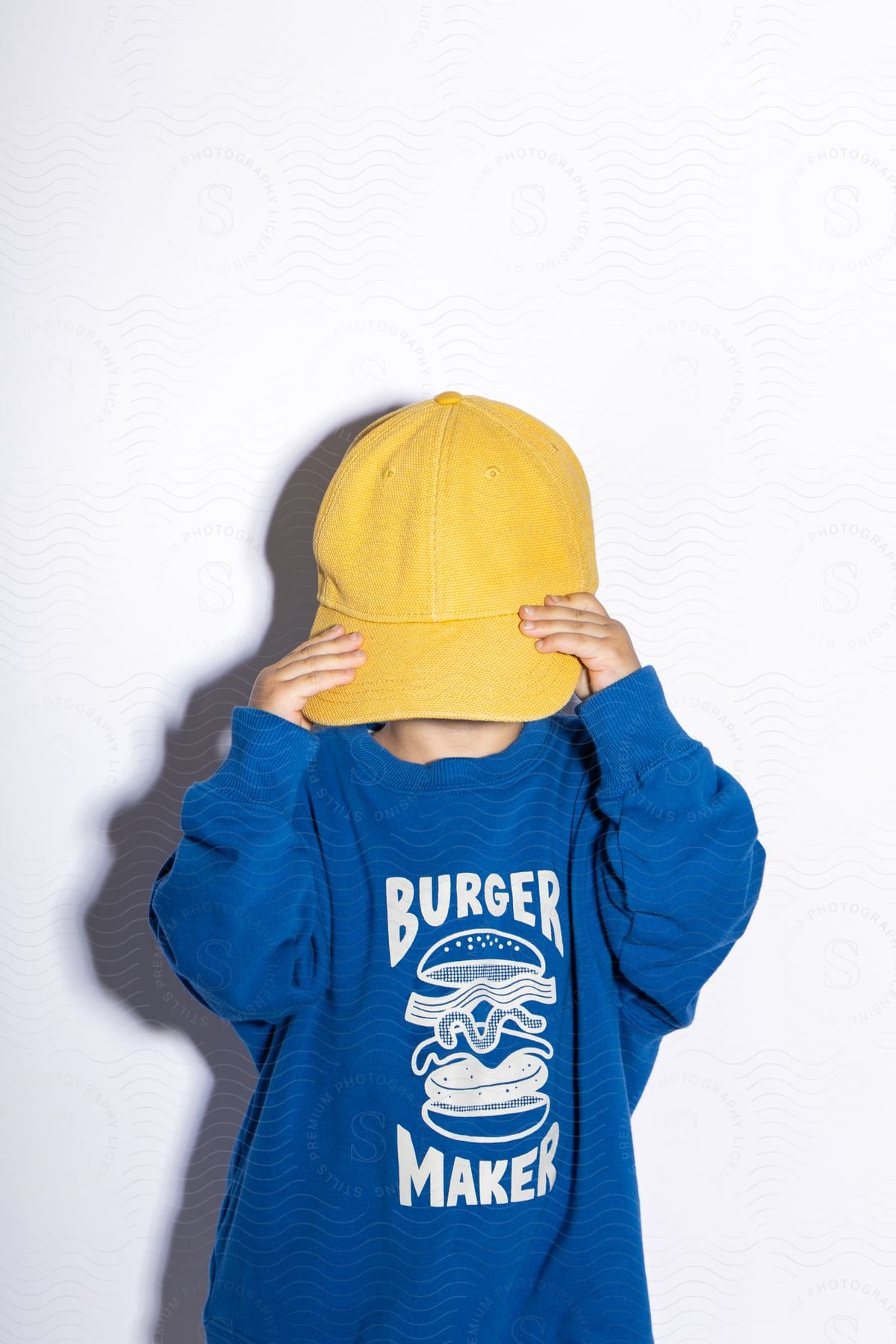 A boy wearing a yellow cap covering his face and a blue hoodie with the words "burger maker" written on it.