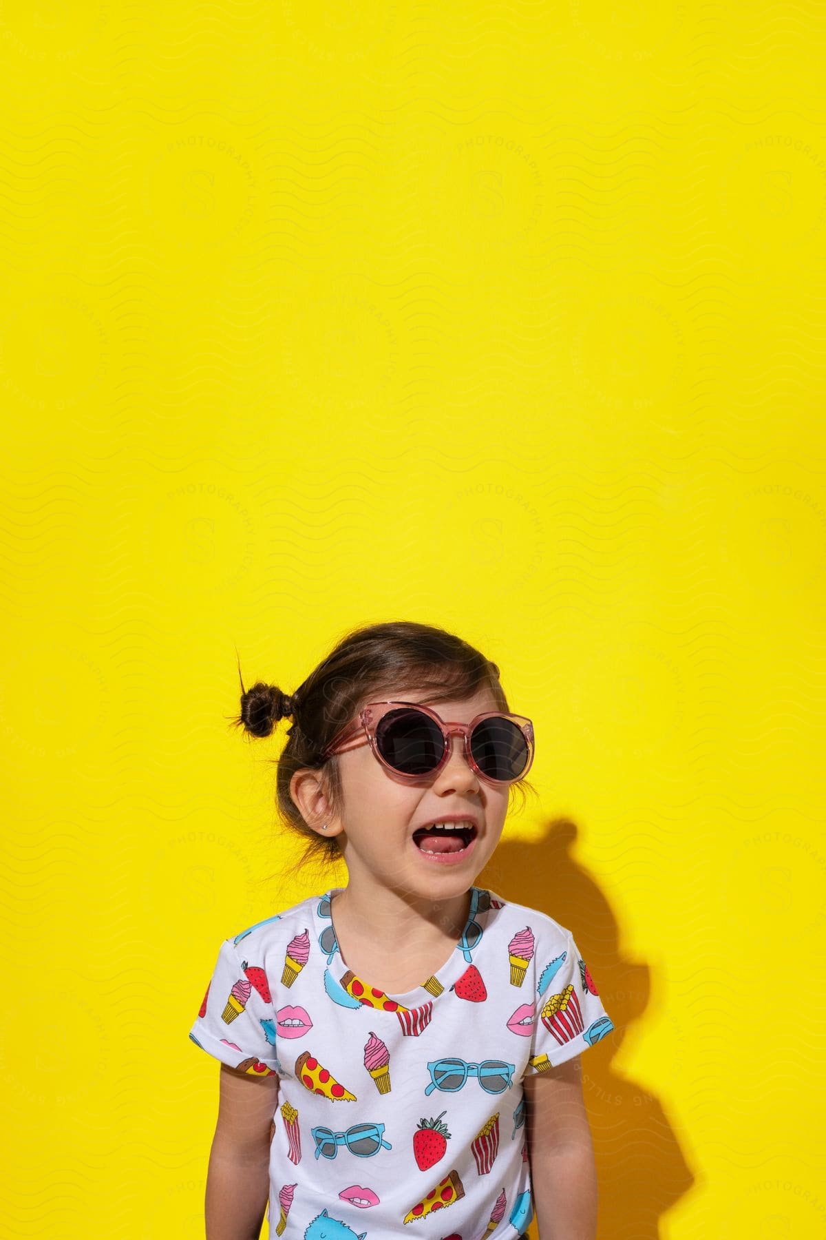 A little girl laughing wearing sunglasses and a shirt patterned with summer treats stands in front of a yellow wall, her shadow merging with the background.