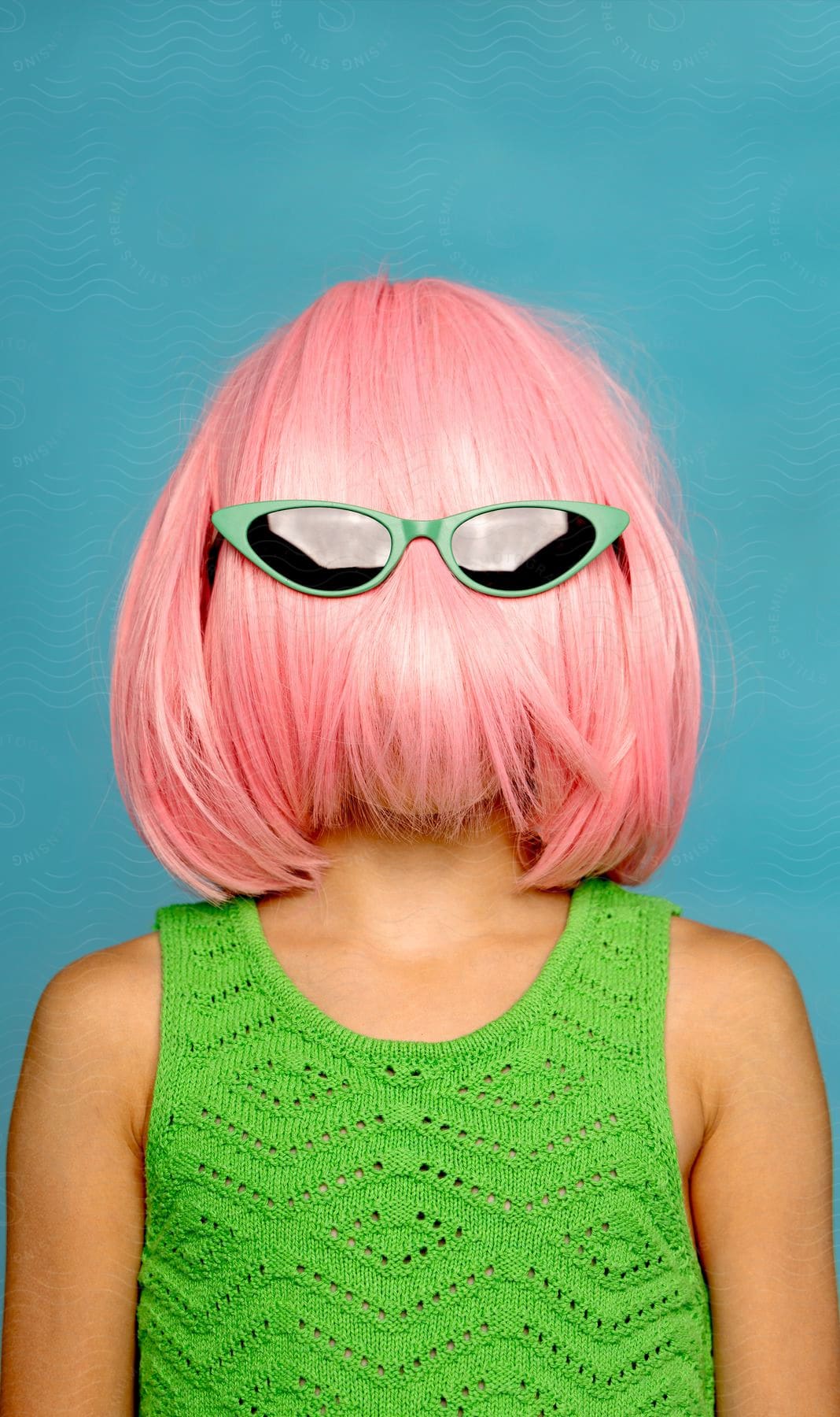 A young girl is posing in green sunglasses and with a pink wig covering her face.