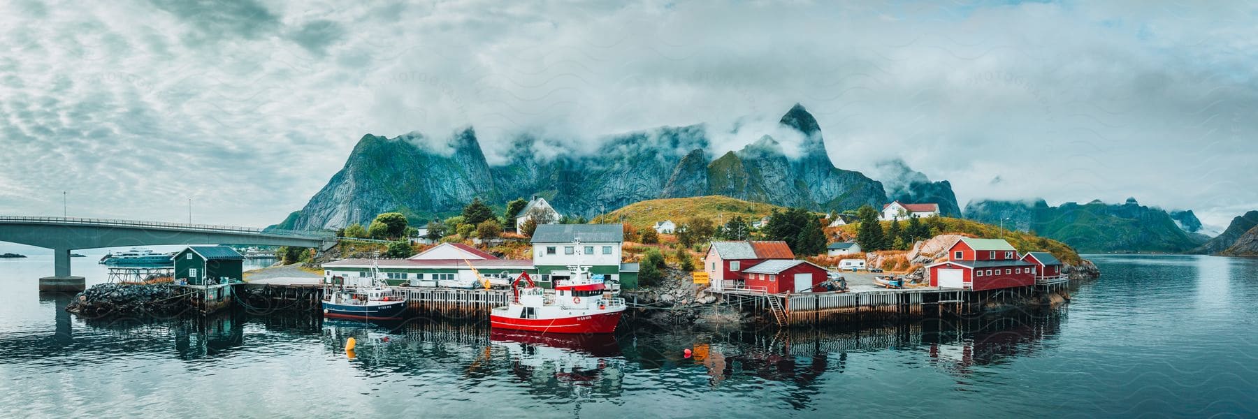 Ships are docked along the coast near houses and buildings as a bridge stretches to the Lofoten Island with mountains in the distance