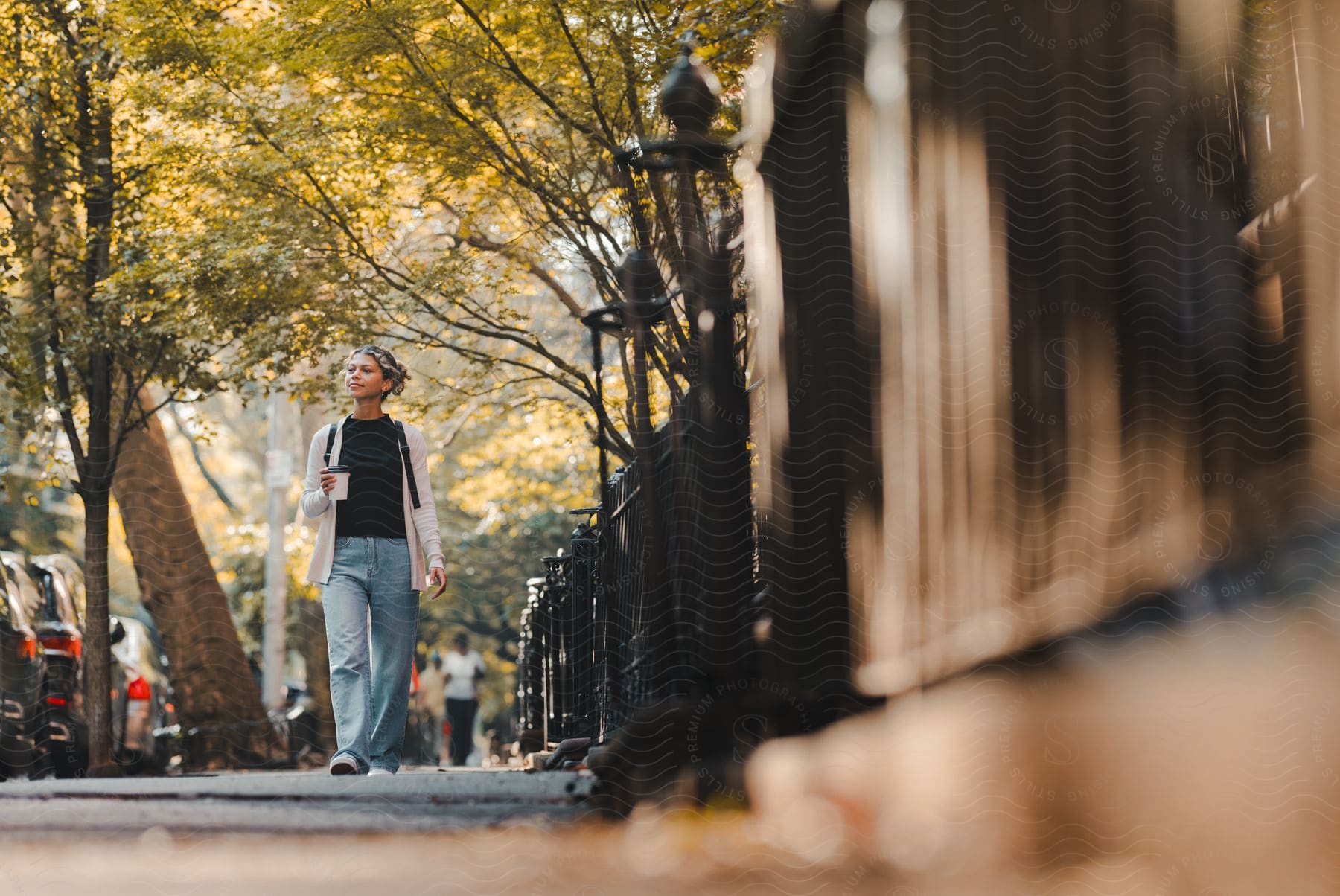 A teenage girl carries a cup as she walks on a busy sidewalk with cars parked on the street