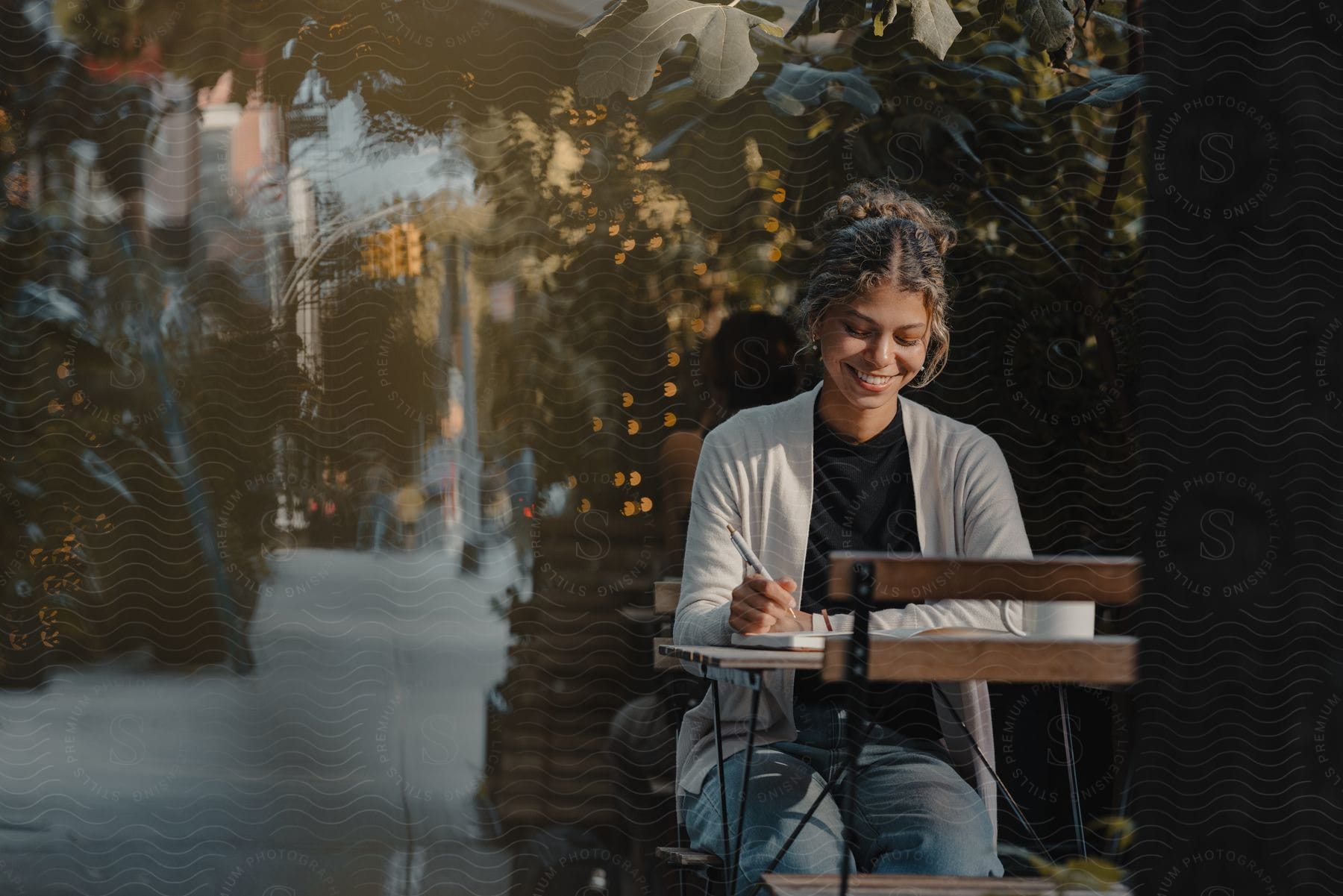 Woman smiling, writing in a notebook at an outdoor cafe table, with green foliage in the background.