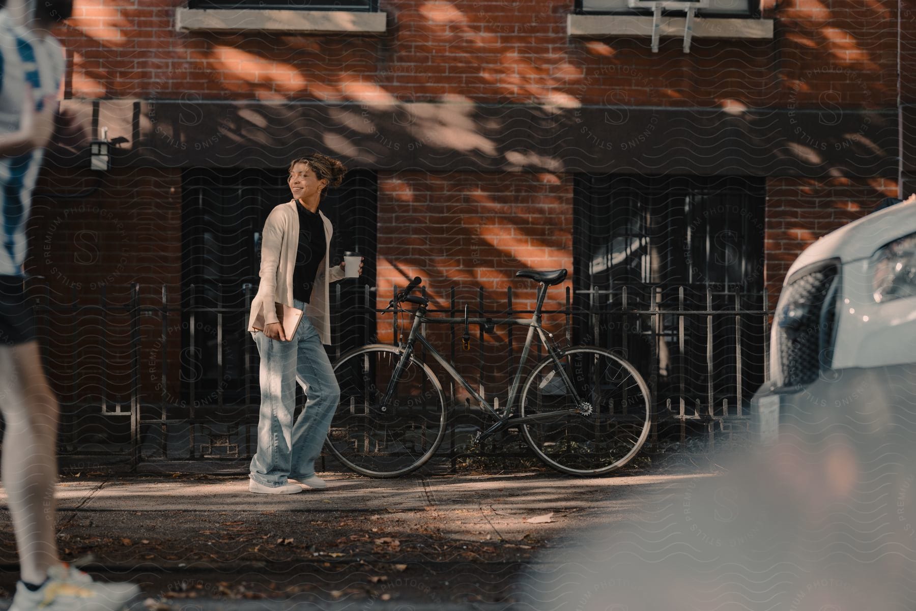 A teenage girl carries a cup and a book as she walks on a sidewalk near a building with a bicycle leaning against a black iron fence