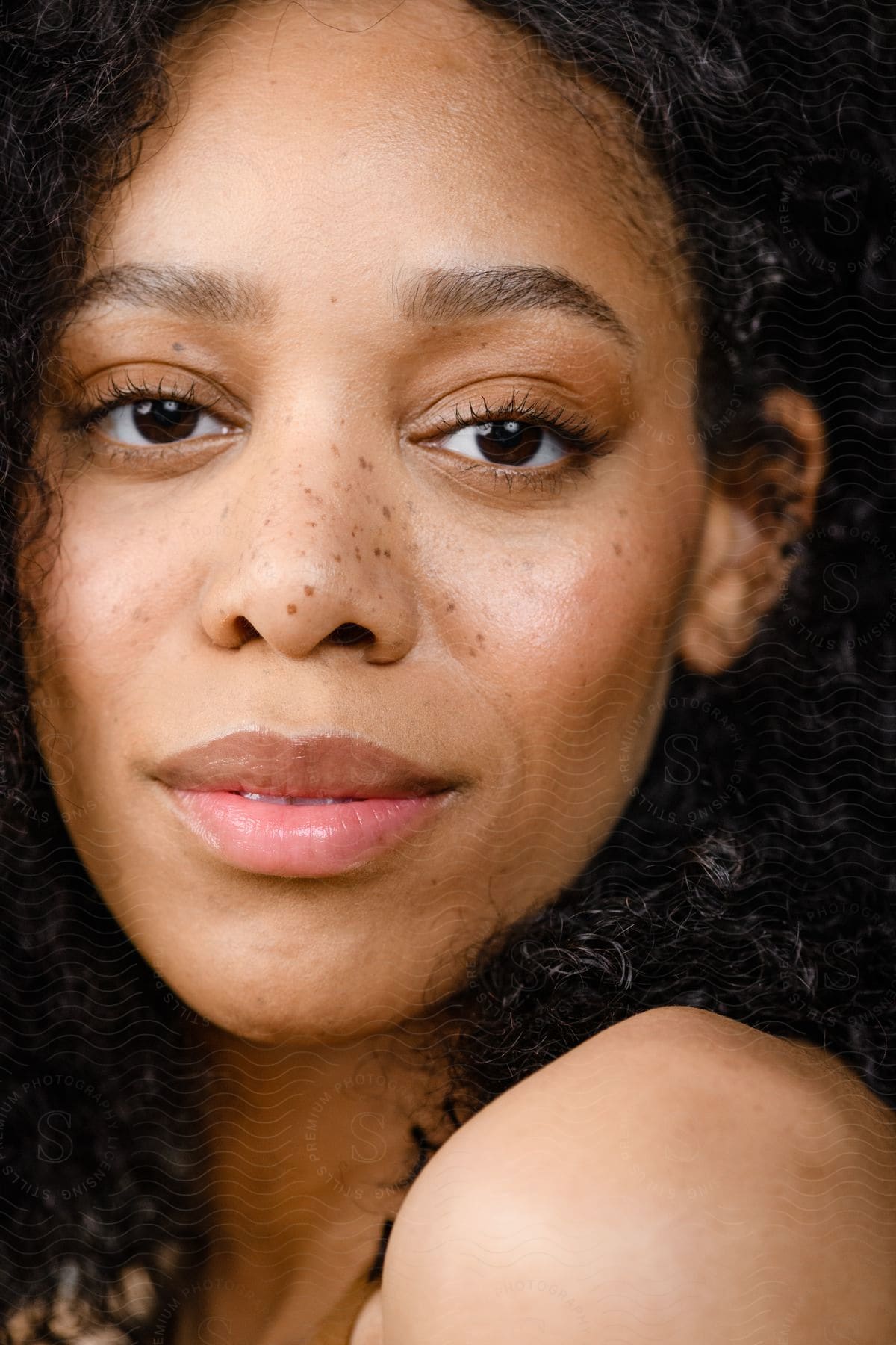 A black woman with freckles, frizzy curly hair gazes neutrally at the camera.