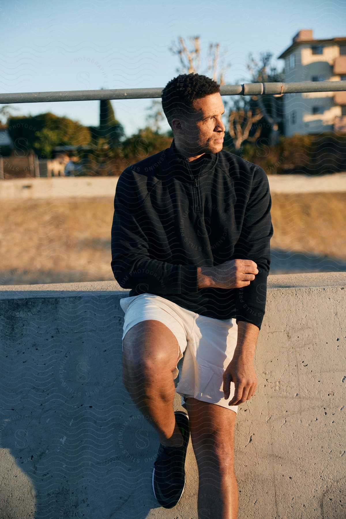 A man in a black coat leans against a small wall as sunlight hits his face.