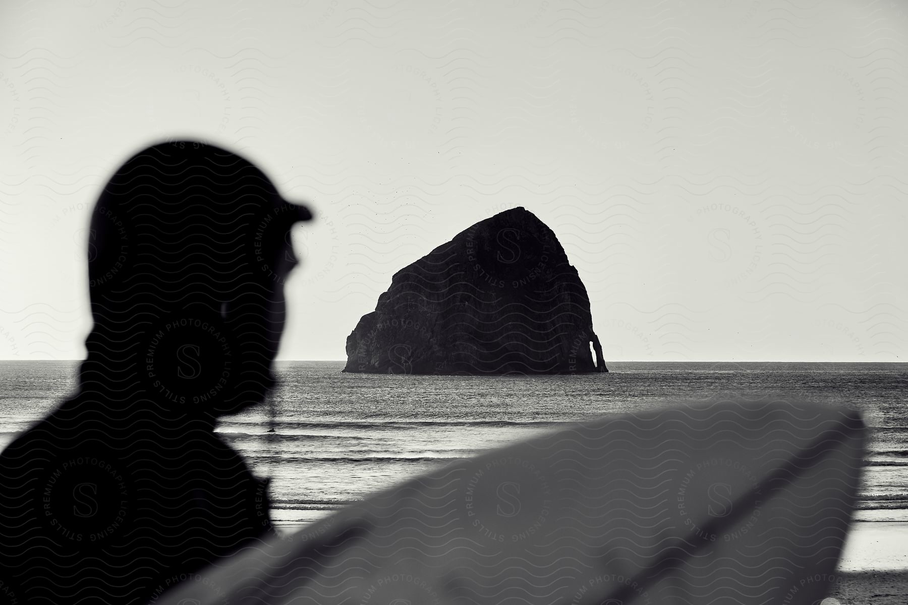 Surfer in silhouette stands on shore looking out to rock island