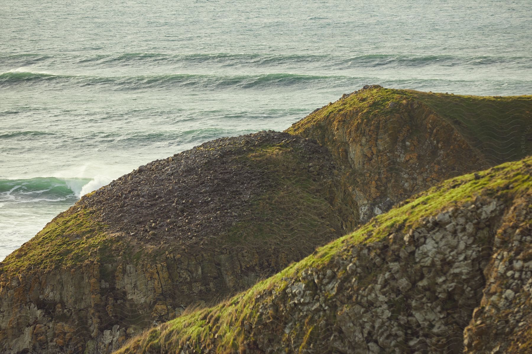 Rocky hills that are covered in green grass are near the ocean which is full of small waves.