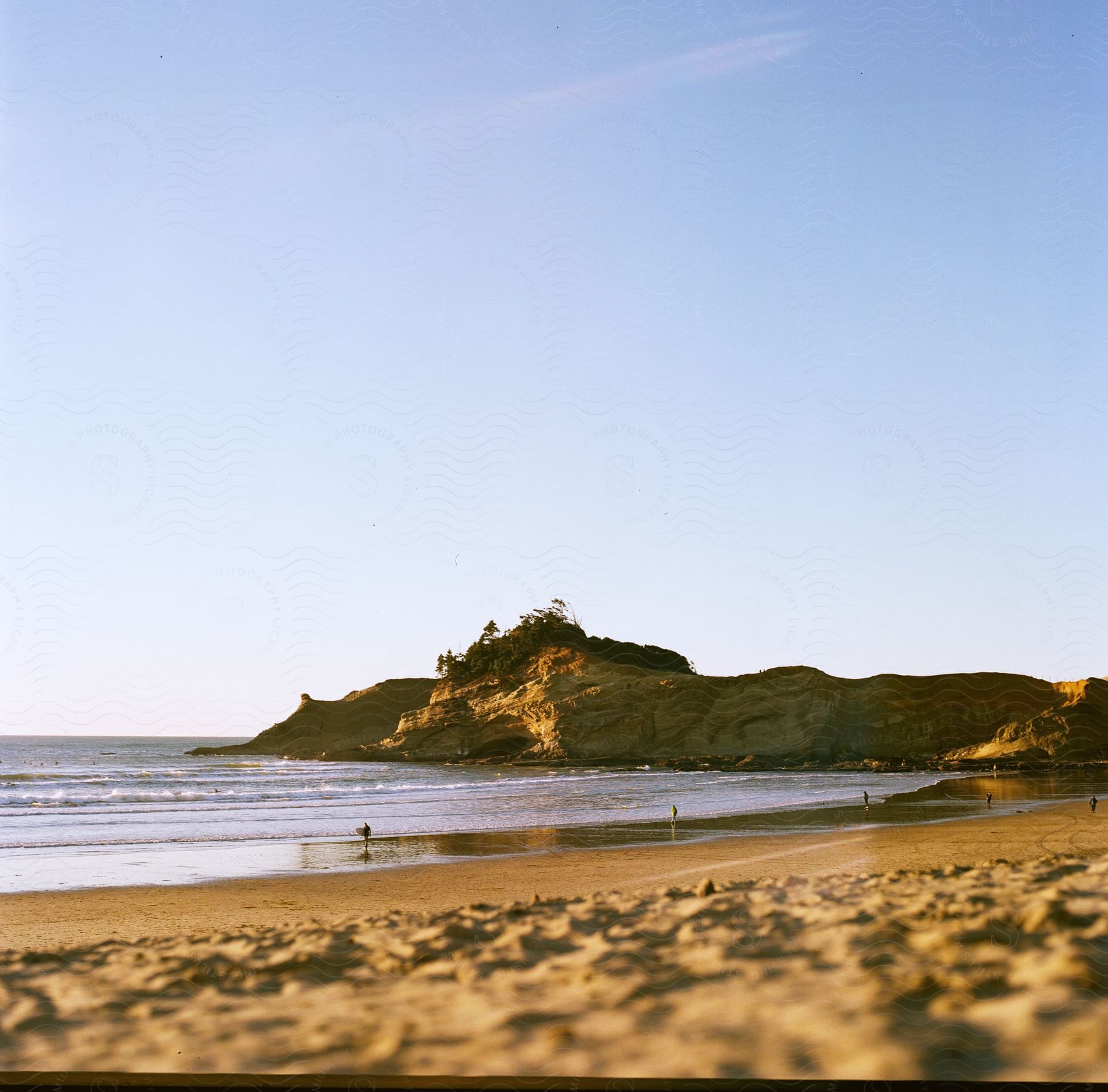 natural landscape shot of a beach next to a promontory with people walking around the sand and one of them carrying a surfboard during day time