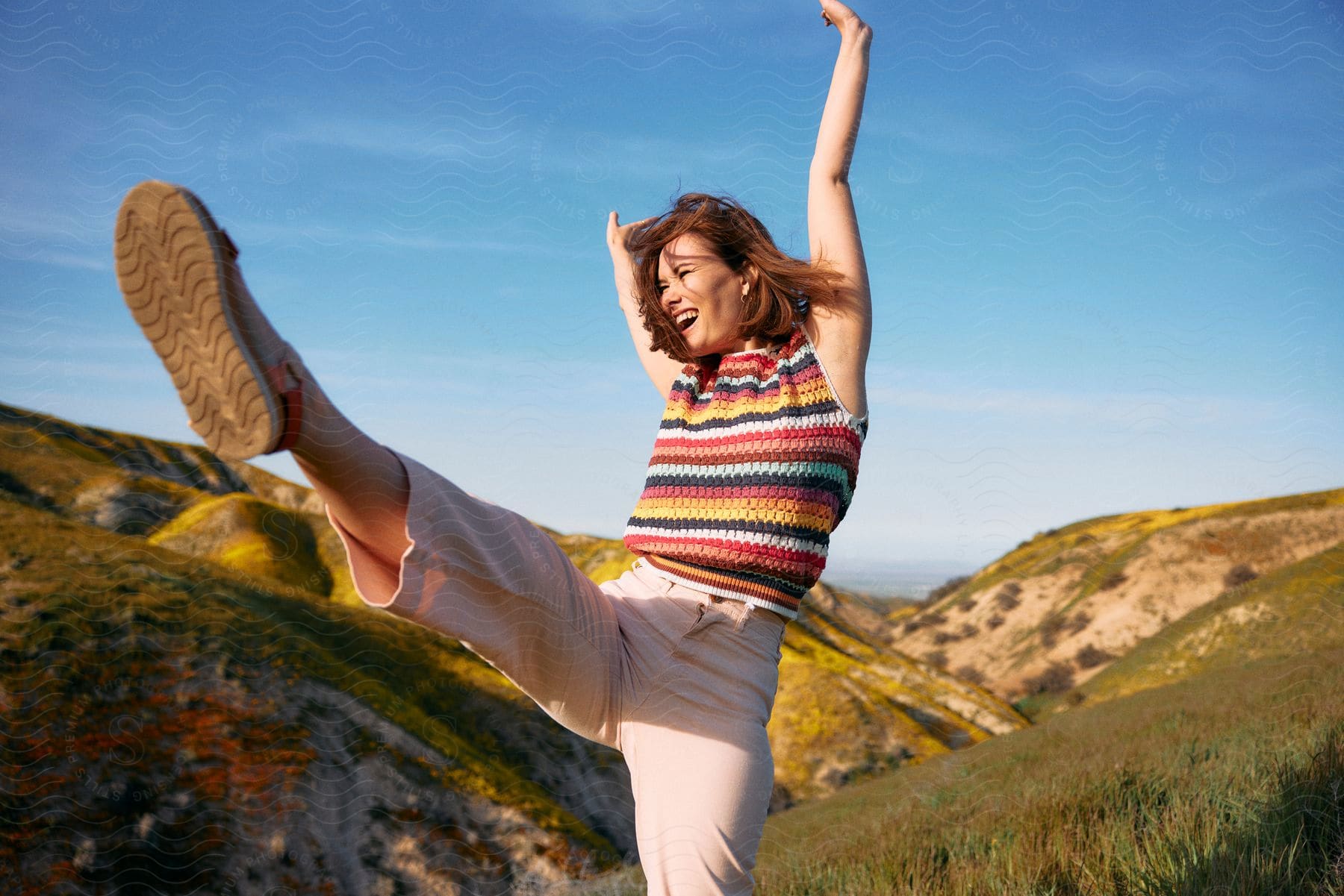 A joyful woman in a multicolored sweater and orange pink pants stands on a grassy hill, arms over head and one leg kicked up in the air, on a sunny day.
