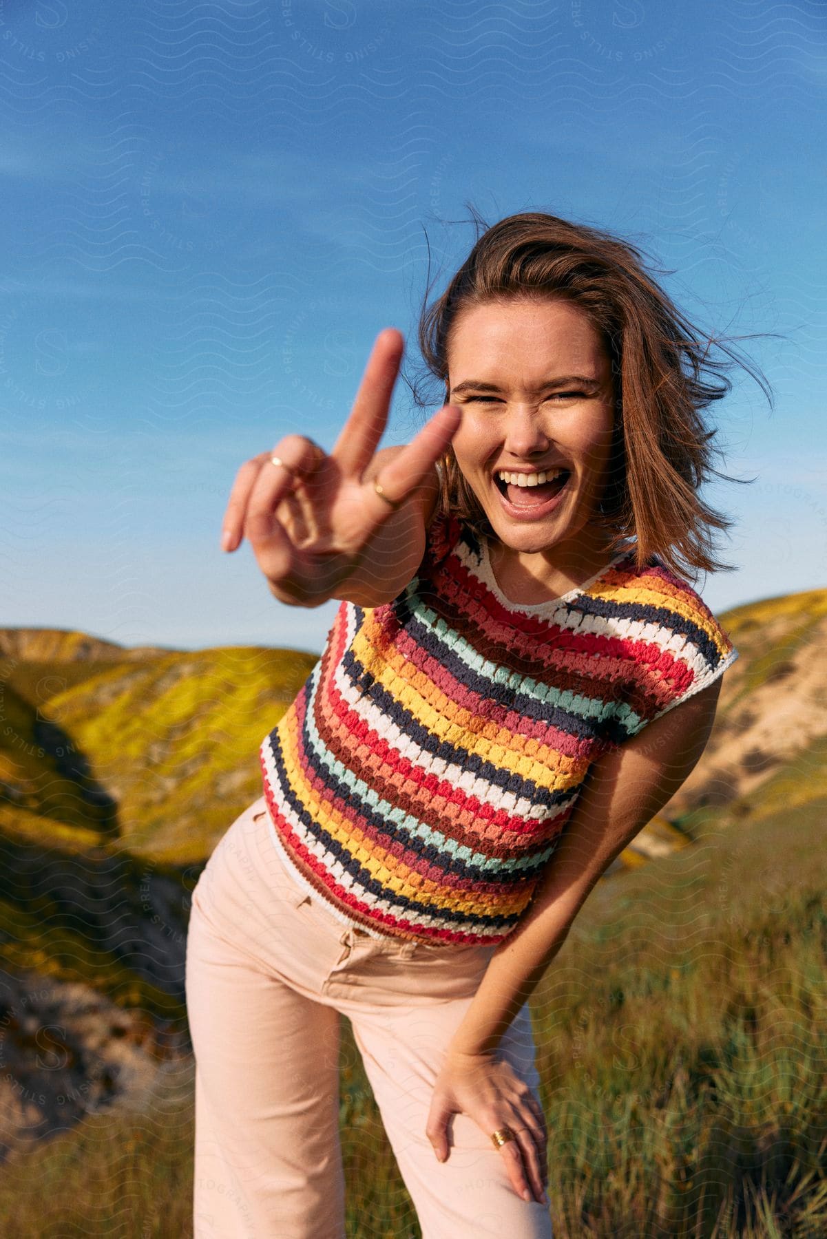 A happy woman in a multicolored sweater and orange pink pants makes a peace sign while standing on a grassy hill on a sunny day.