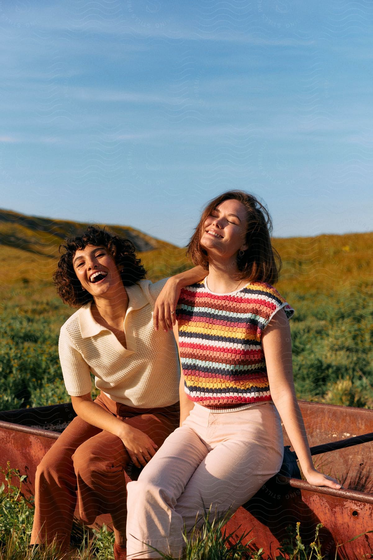 Two young women are smiling while sitting on a rusted car frame in a remote area.