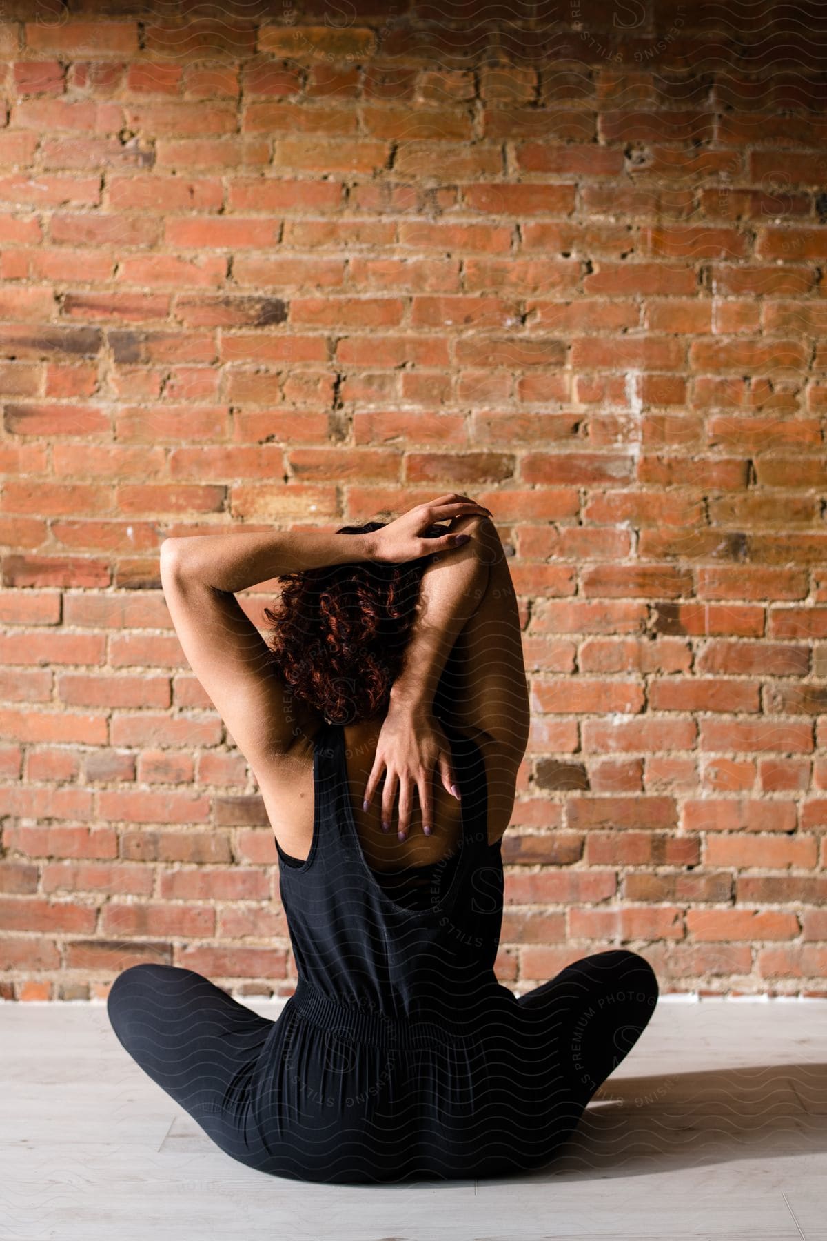 A woman is sitting on the floor facing a wall with her arms in the air and one hand stretched to the middle of her back and the other hand on her elbow