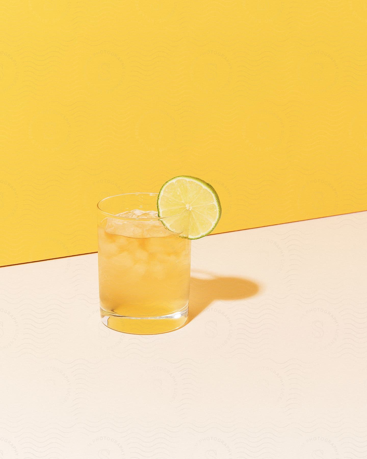 iced yellow beverage served on a glass with a slice of lime over a white table next to a yellow wall
