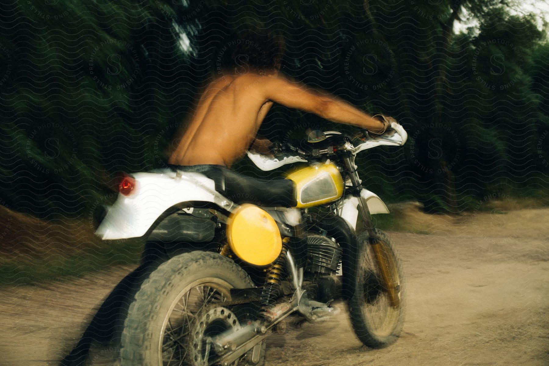 A shirtless man pushes a motorcycle along a dirt road.