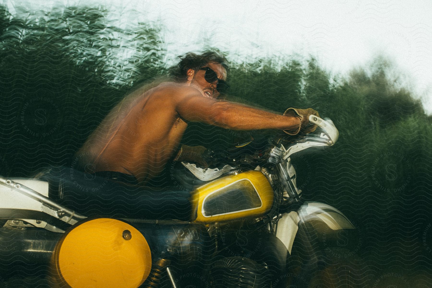 A bare chested man wearing sunglasses turns his head as he rides a motorcycle near the woods