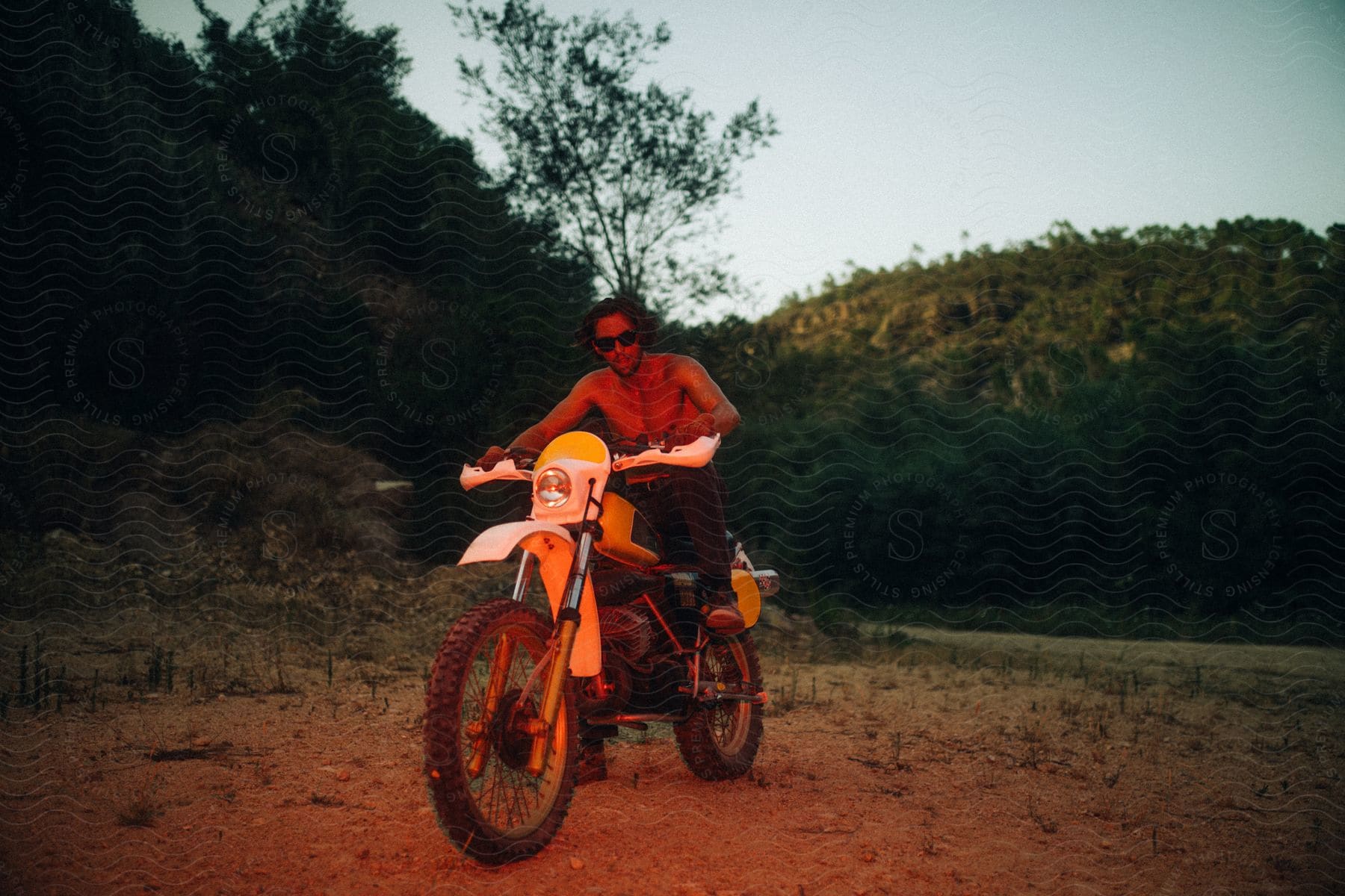 Topless man with sunglasses sits on dirt bike on dirt road