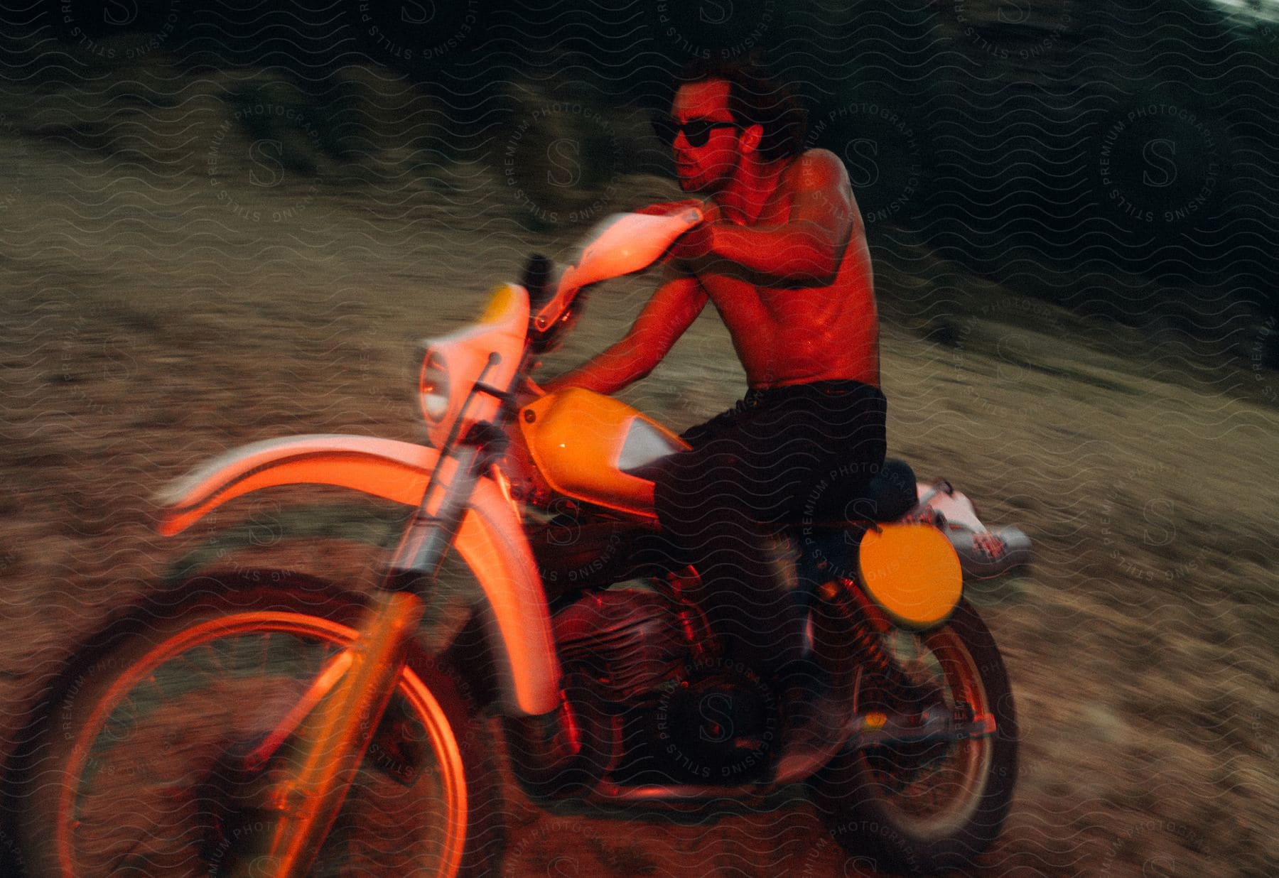 A bare chested man wearing sunglasses is riding a motorcycle on a hillside