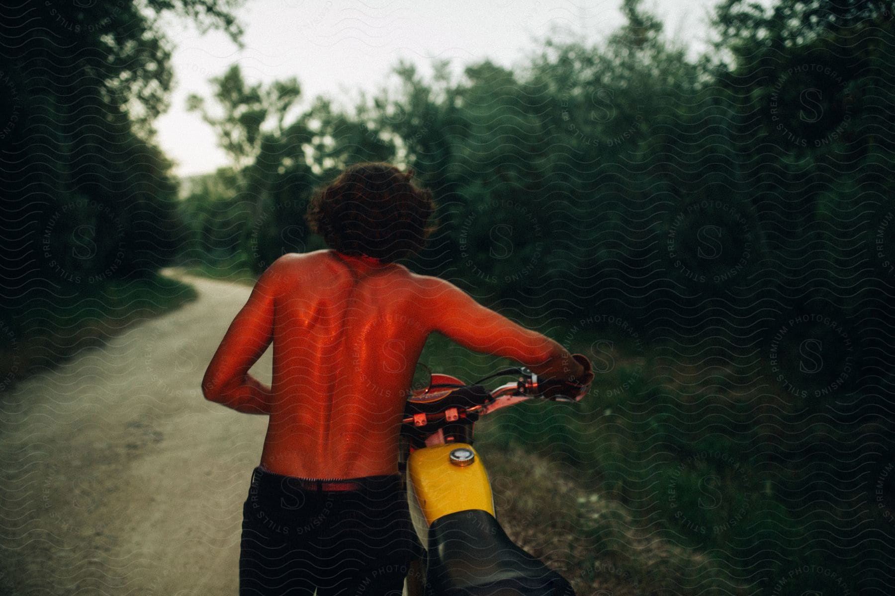 A man wearing no shirt is pushing a motorcycle on a road through the woods