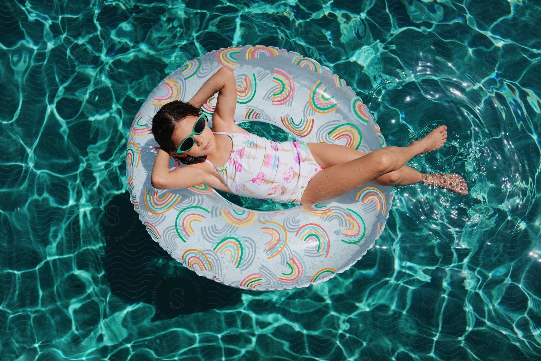 A rainbow-clad girl floats on a matching inflatable ring in a pool, her reflection dancing on the water below, green sunglasses shielding her eyes from the sun.