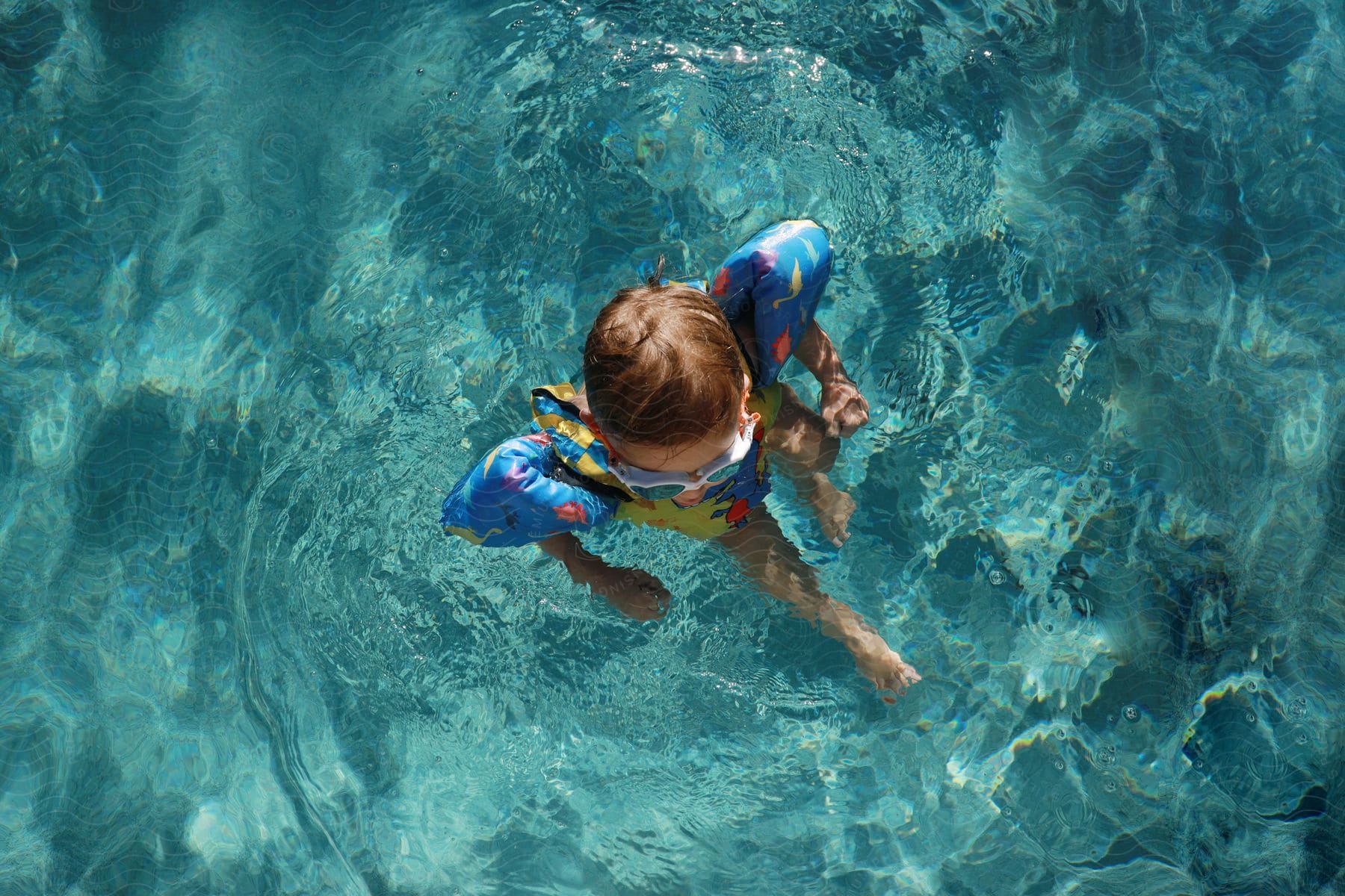 A young child wearing arm floats is sitting in a shallow pool or body of water.