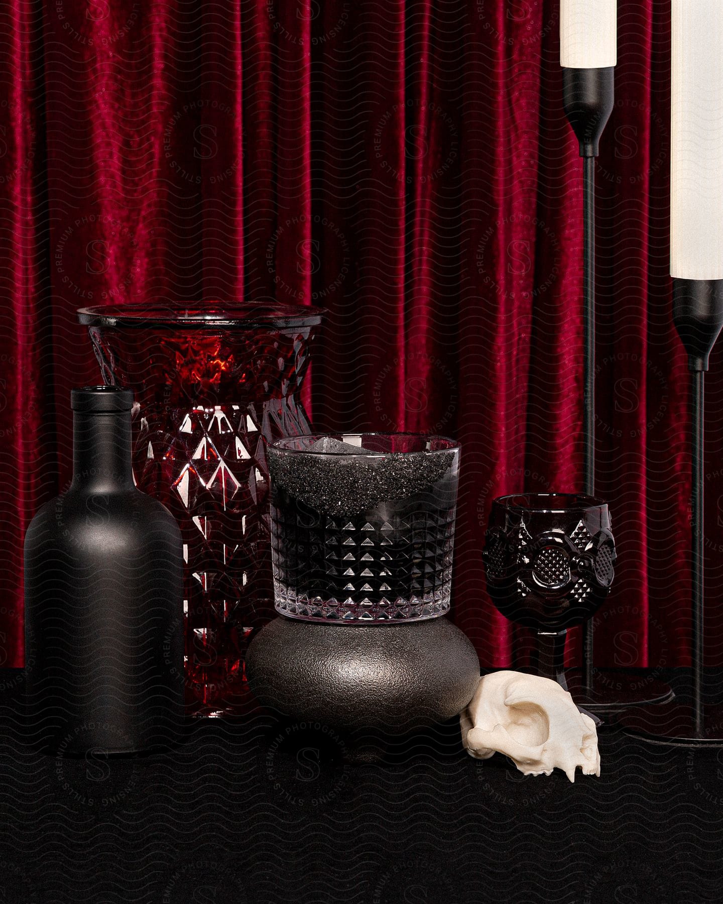 Red and black vessels and a skull and candles sit near red curtains.