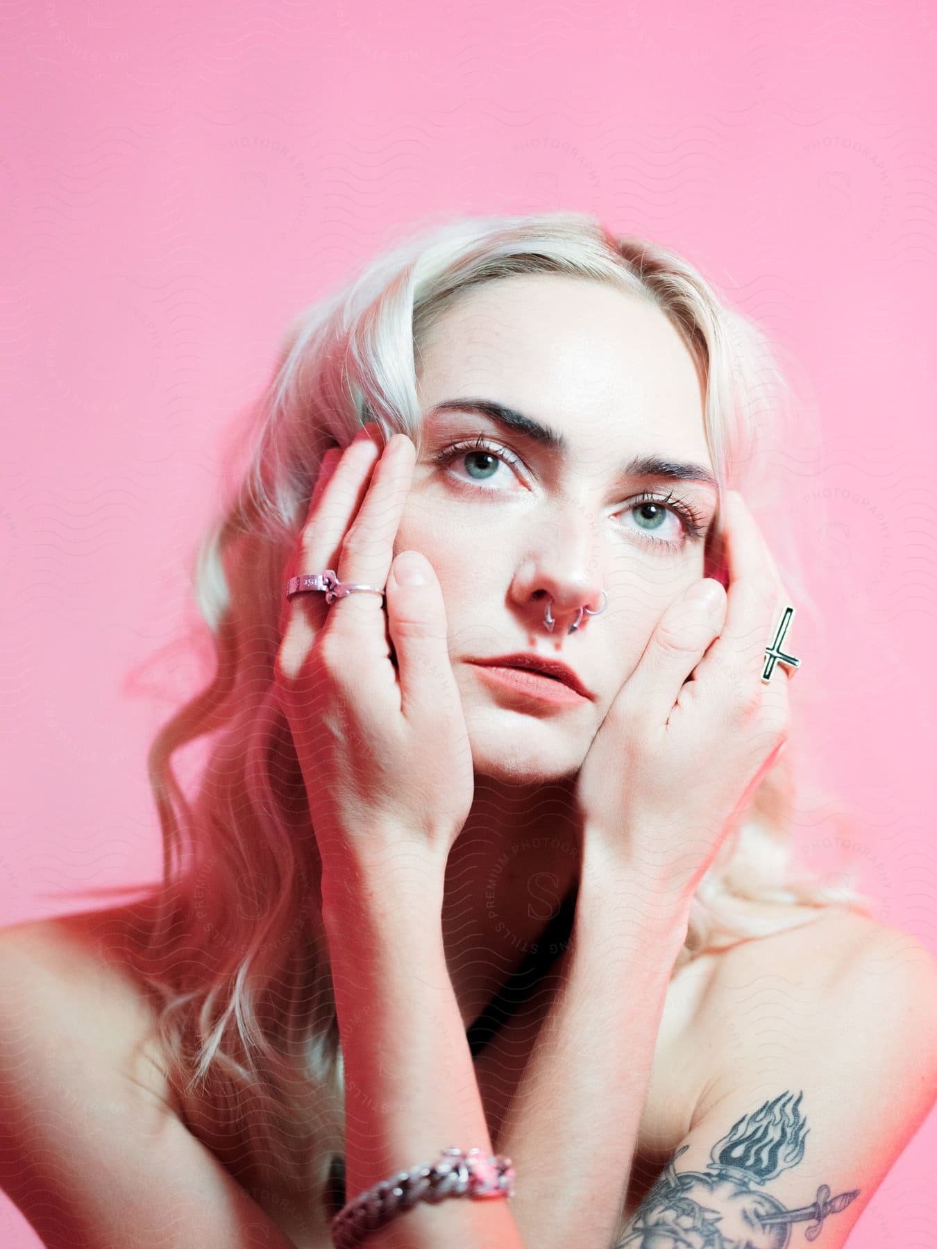 Portrait of a young blonde woman with her hands on her face wearing rings, jewelry, and a tattoo on her arm.