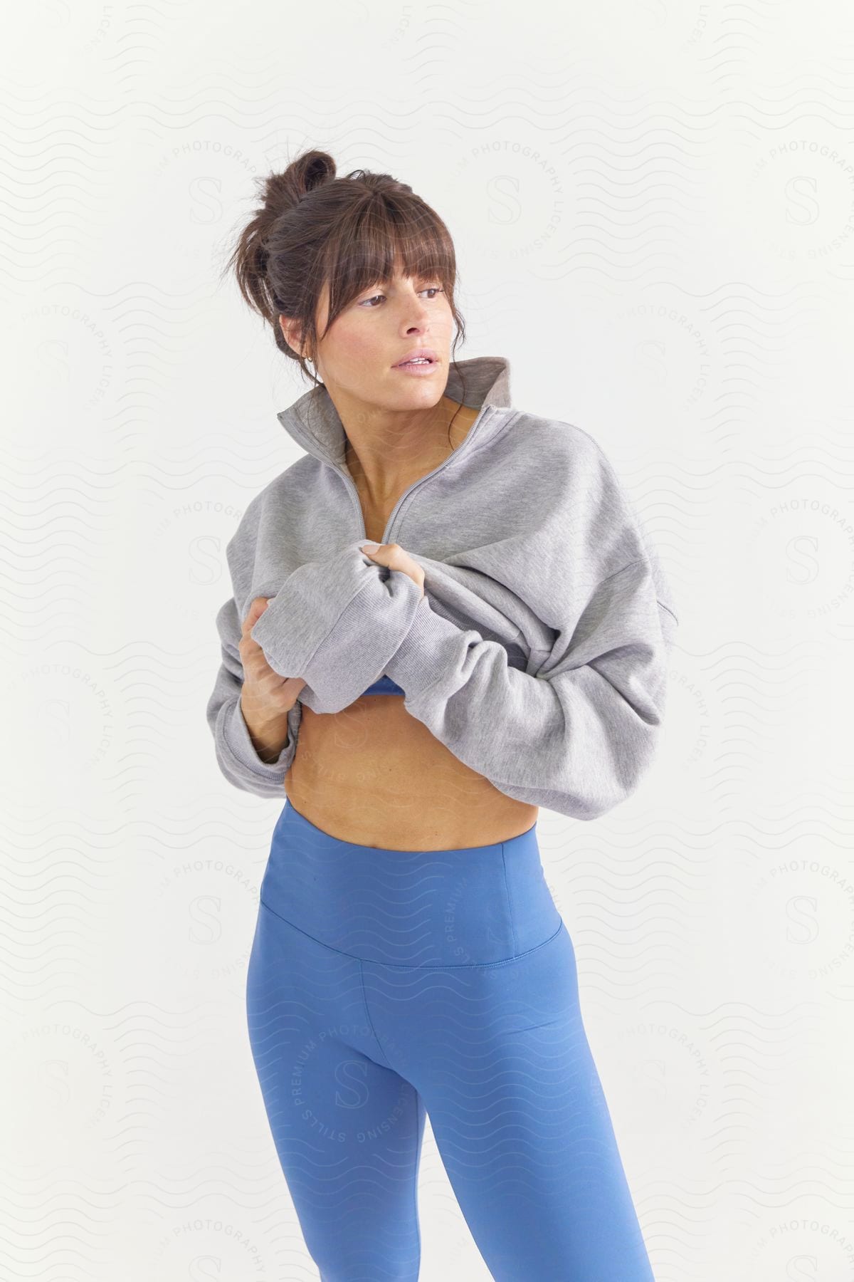 A woman in grey sweatshirt and blue yoga leggings lifts her sweatshirt to reveal her bare abs and blue bra.