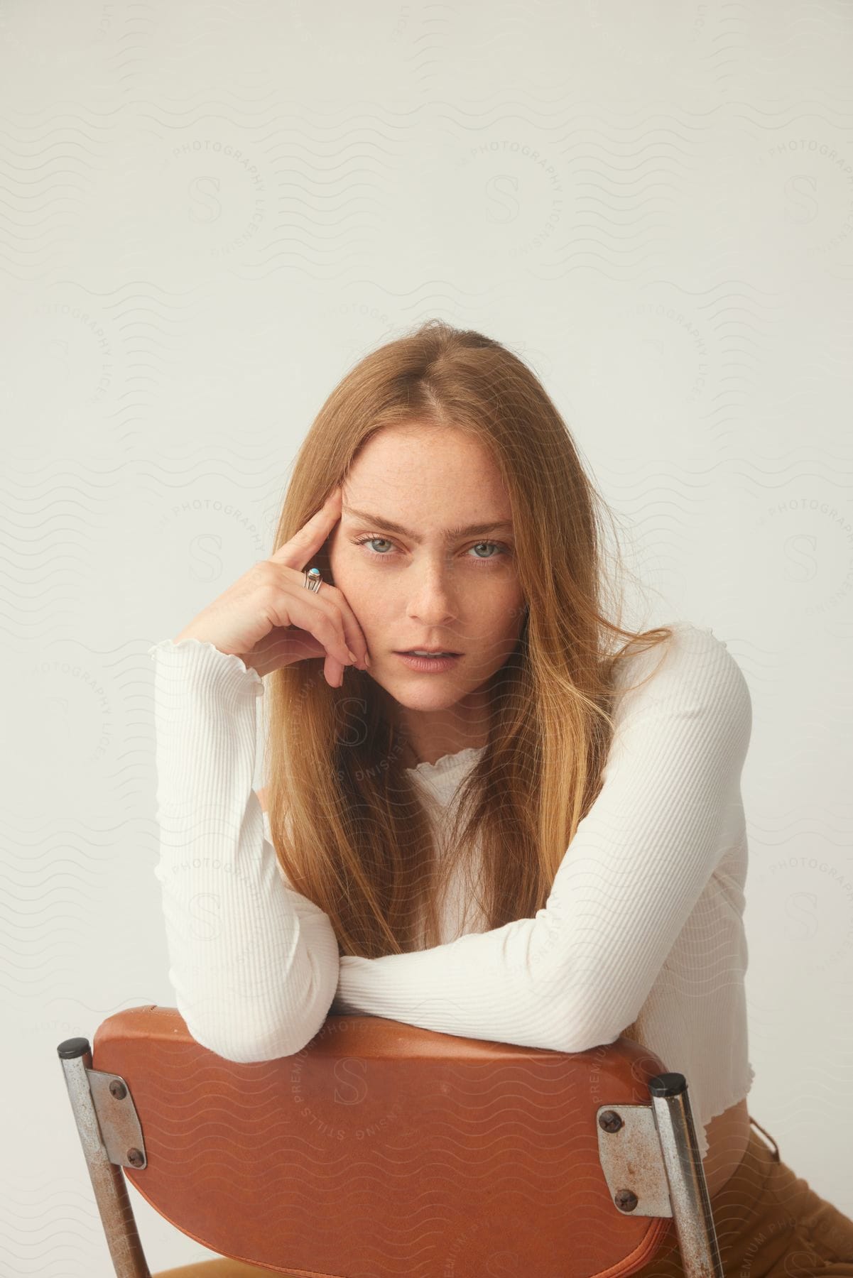 A woman with long blonde hair is sitting on a chair with her elbows on the backrest and a hand on her face as she looks ahead