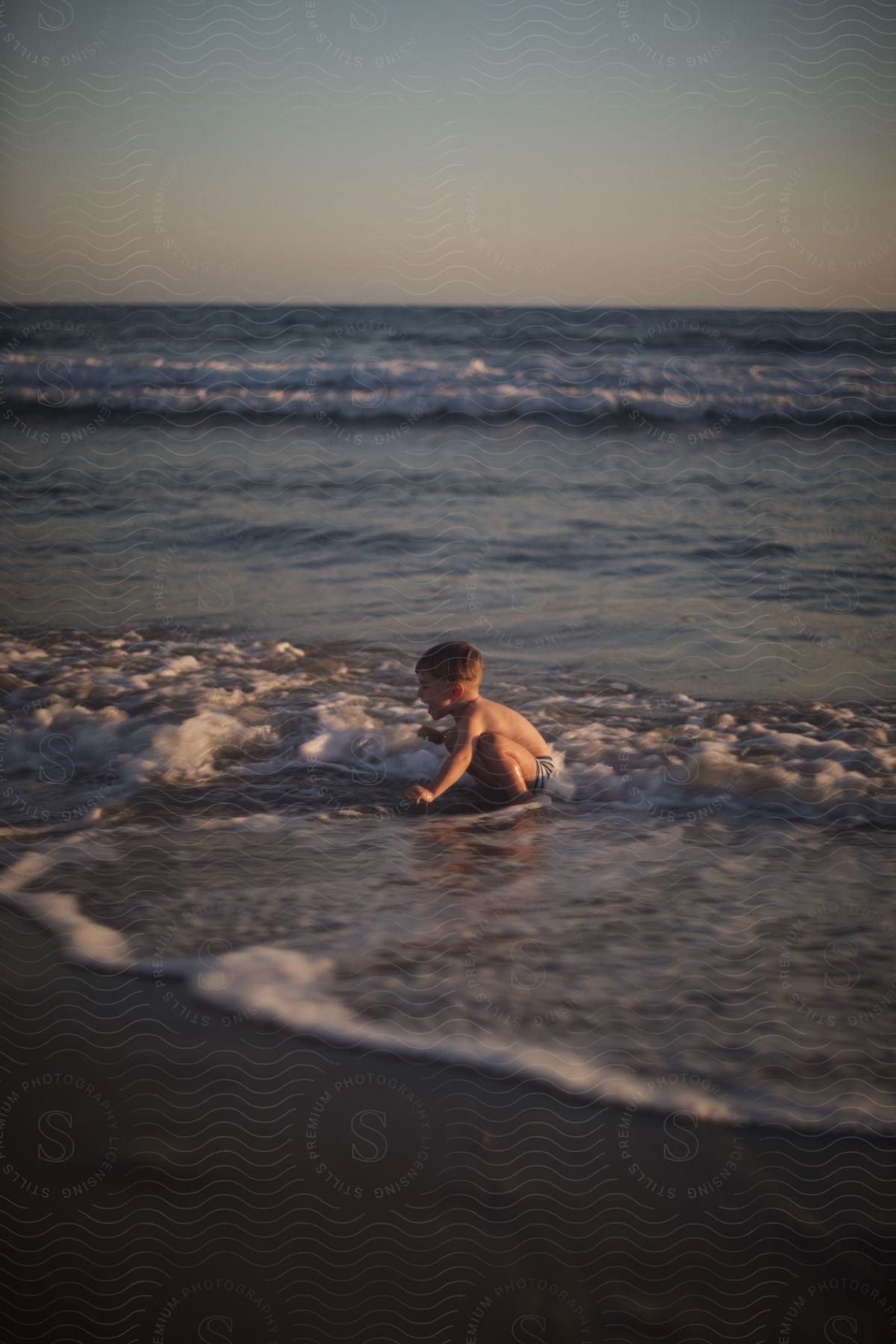 Young boy squats in the shallow waves lapping up on the beach.