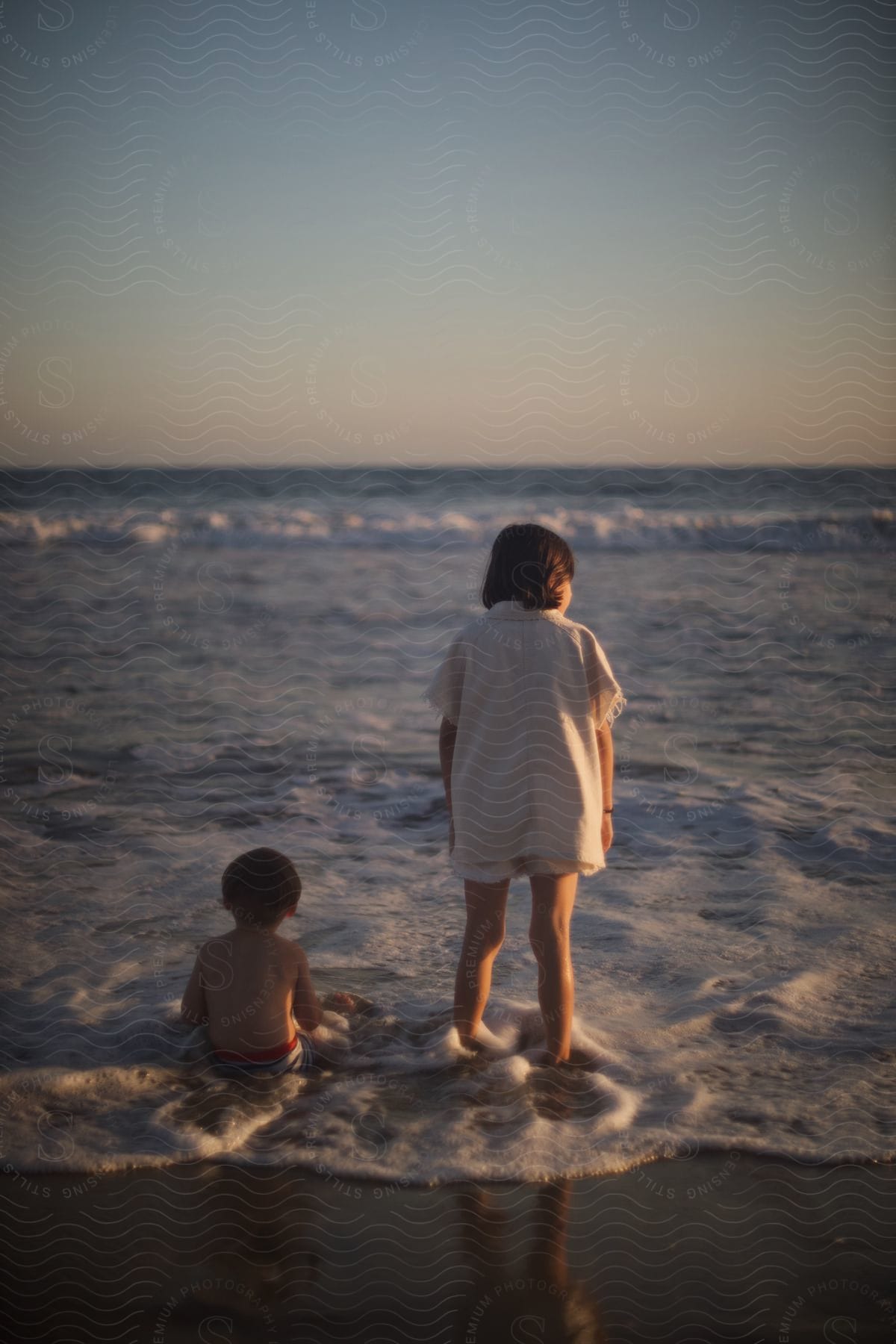 Two young people on a beach at sunset, one is sitting and the other is standing.