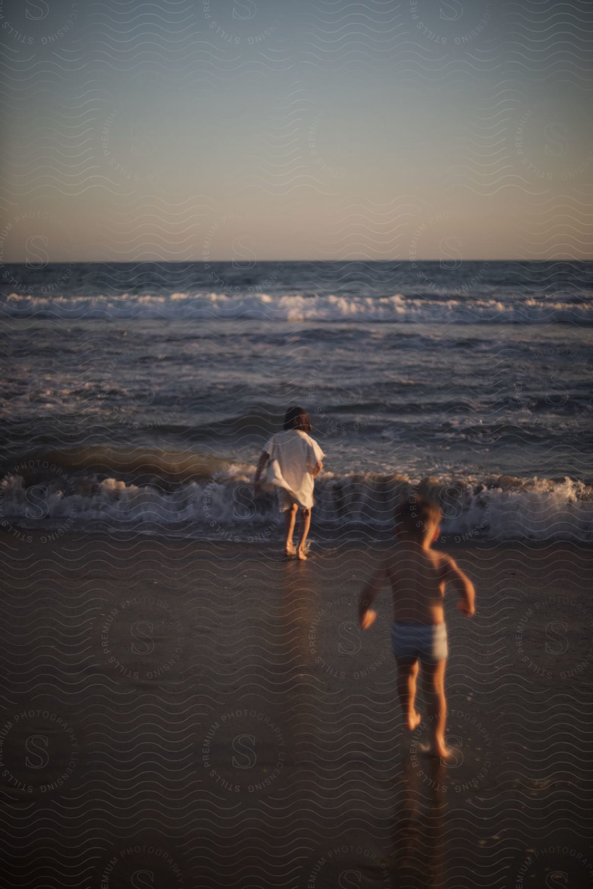 Siblings run toward surf on beach as red tints the horizon.