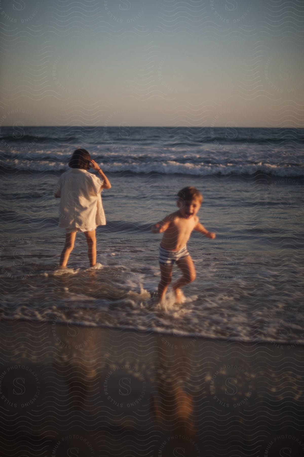 A girl and a male toddler are wading in ocean water on the beach on a clear day.